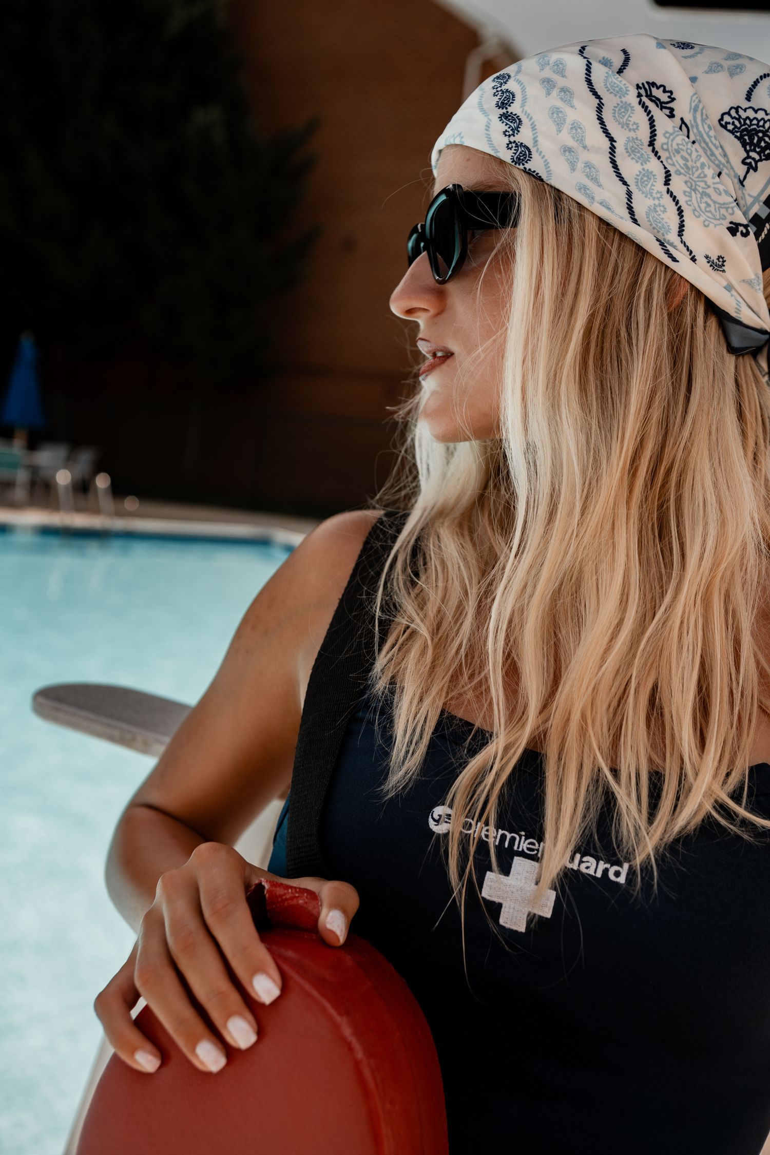 Sunlit profile view of someone in lifeguard attire wearing a patterned hat near a pool.