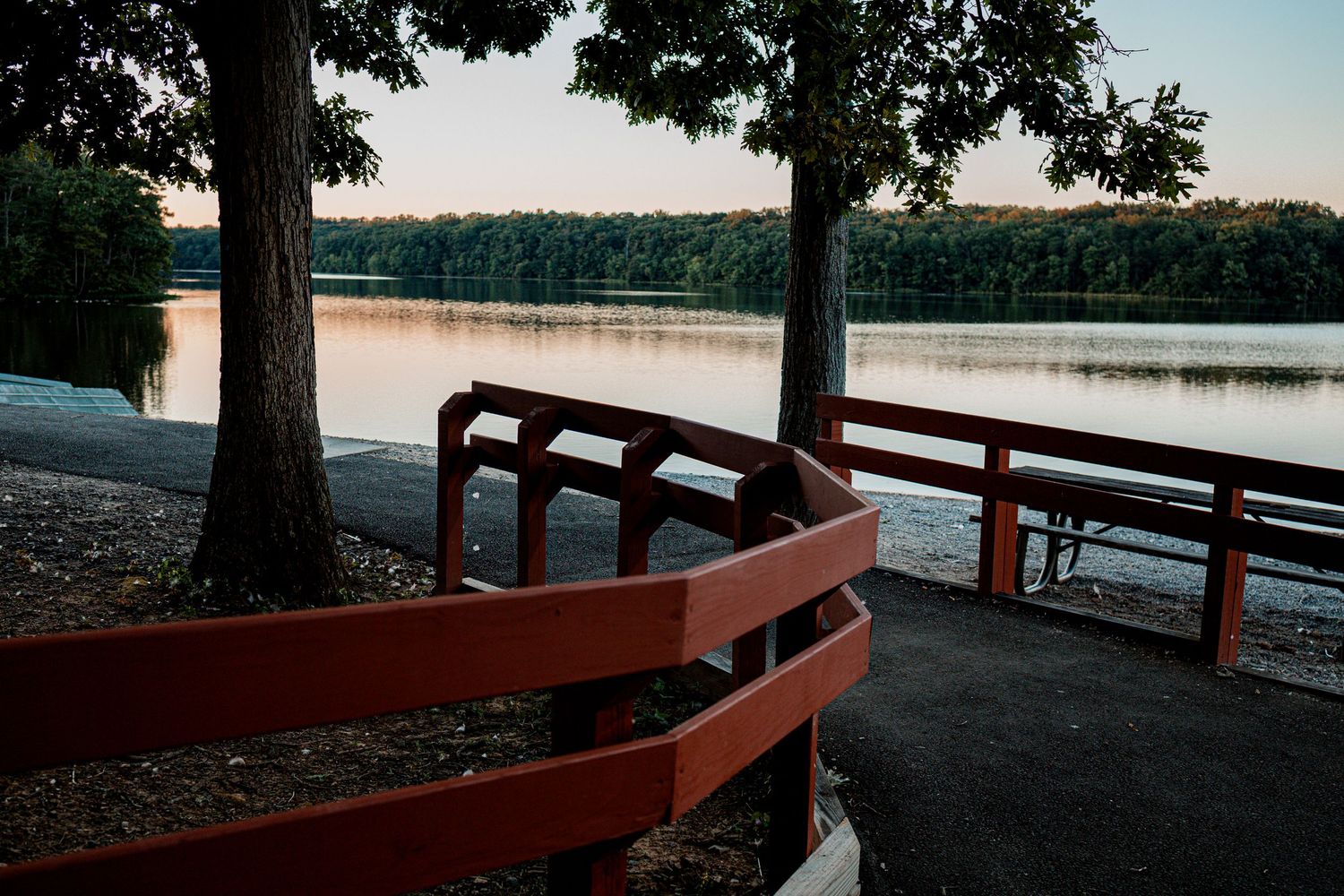 A curved wooden boardwalk along a serene lakefront with trees and water views at sunset.