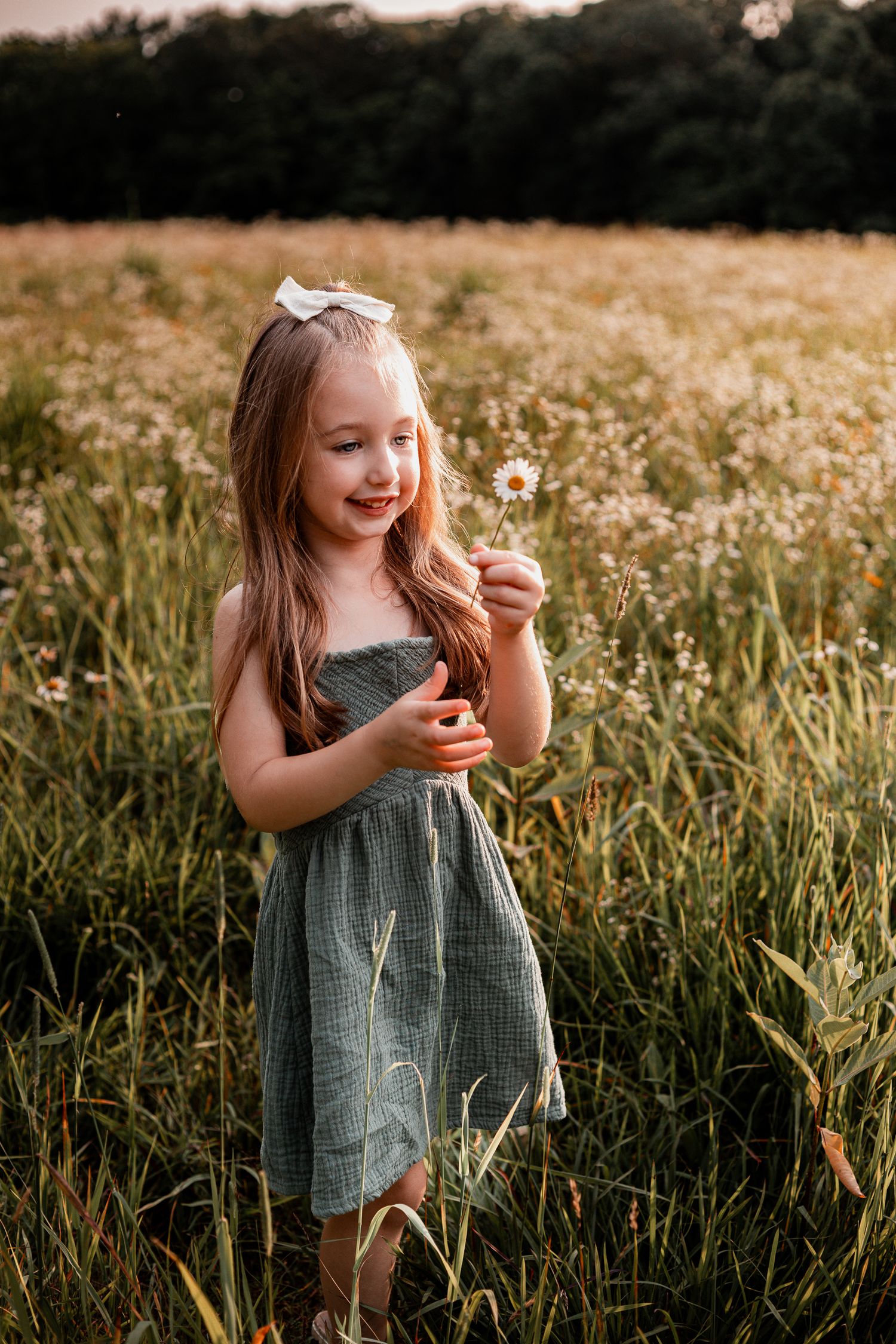 A child in a gray dress laughs while holding wildflowers in a summer meadow at sunset.