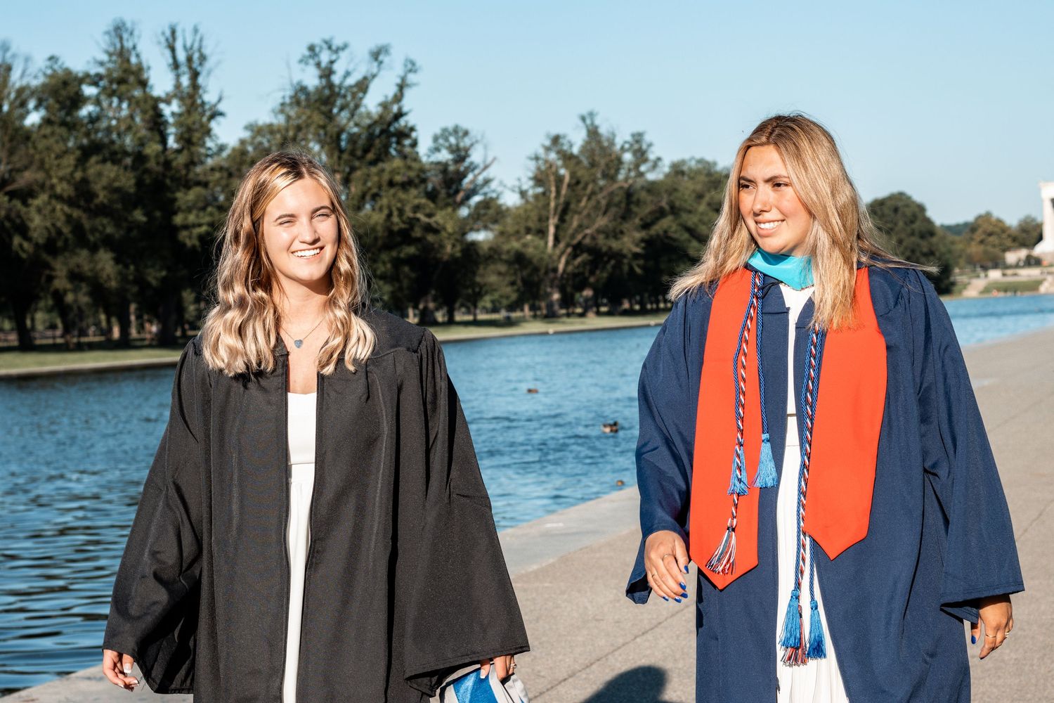 Graduates in academic regalia taking celebratory photos by a lakeside on a sunny day.