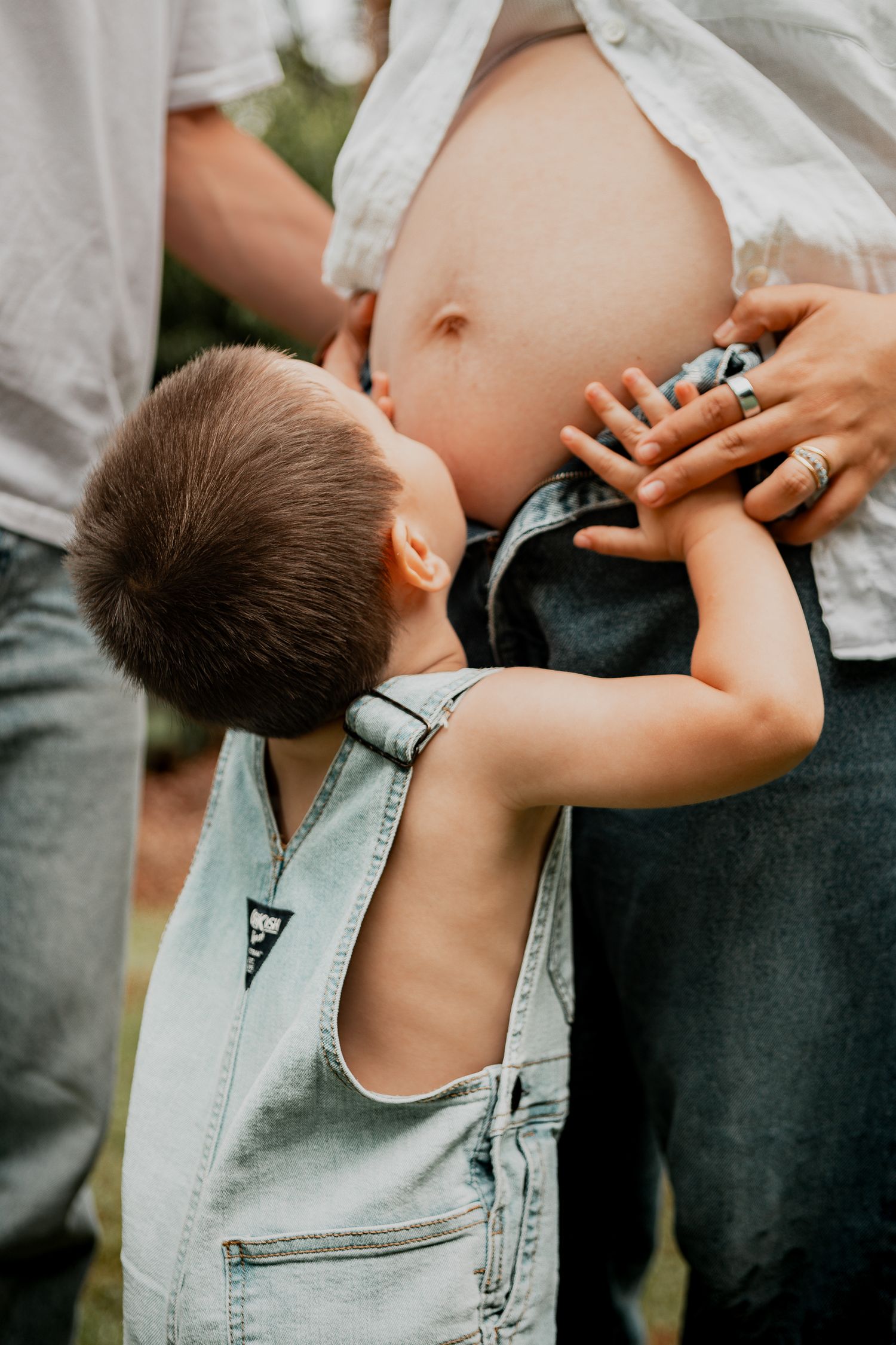 A small child in denim overalls touches a pregnant belly with curiosity and wonder.