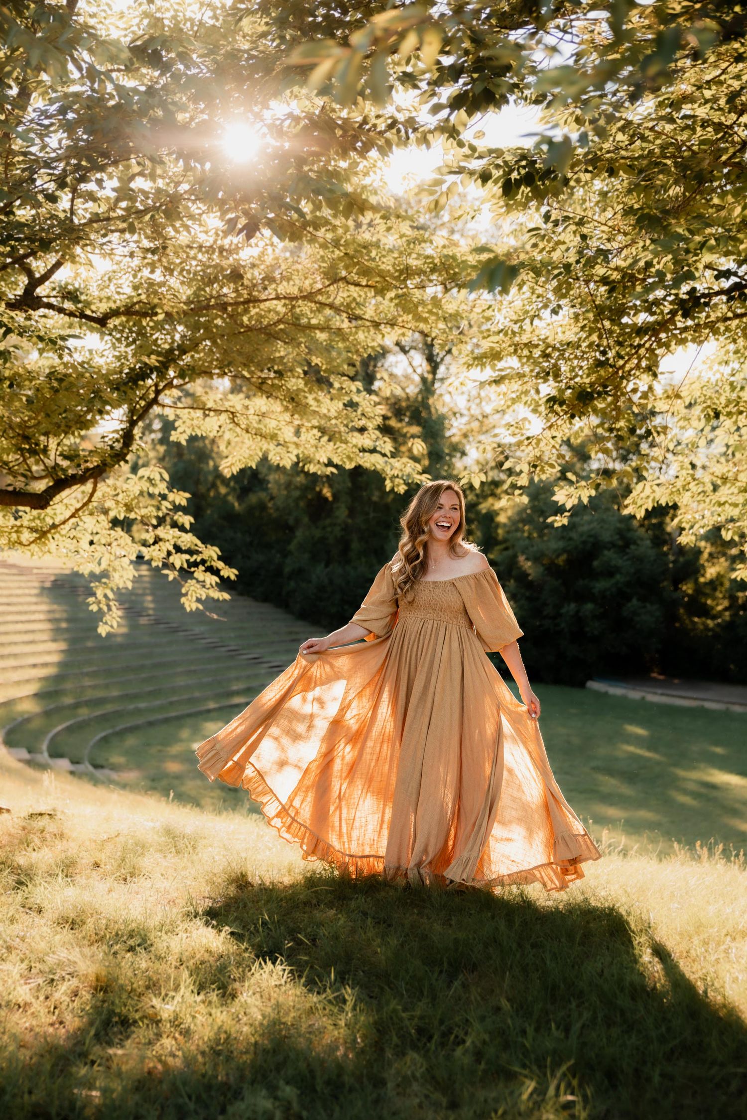 Woman in golden off-shoulder gown twirling in sunlit park under lush green trees during golden hour.