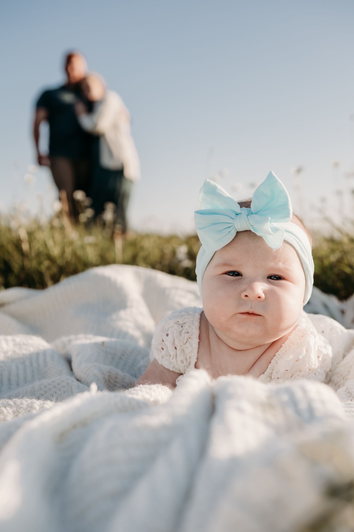 A small infant wearing a blue bow lies on a white blanket outdoors.