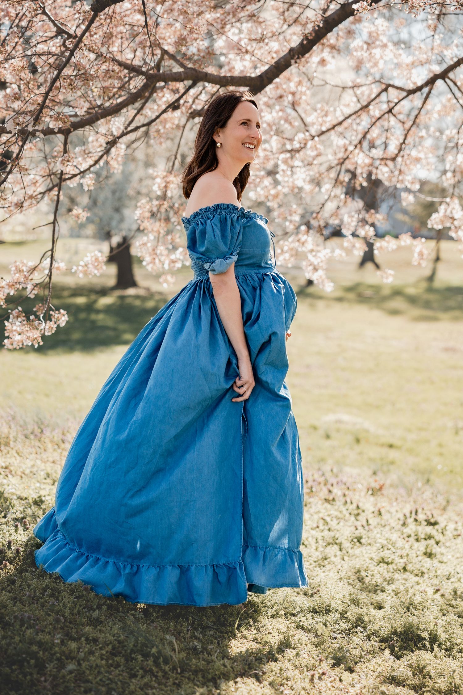 Woman in blue off-shoulder ball gown standing under cherry blossom trees in a sunlit garden. Maternity Cherry Blossom Session Washington D.C
