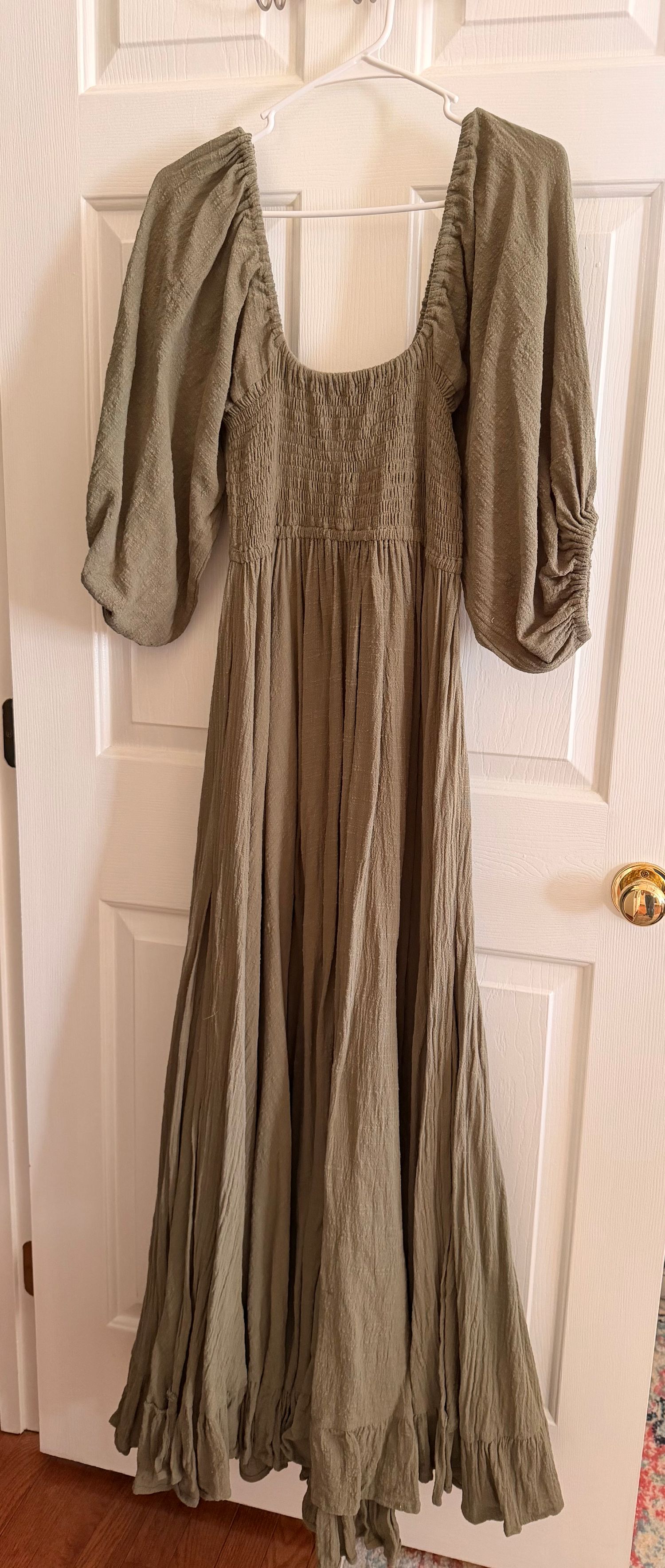 Olive green maxi dress with puffed sleeves, smocked bodice, and flowy ruffled skirt hanging on a white door.