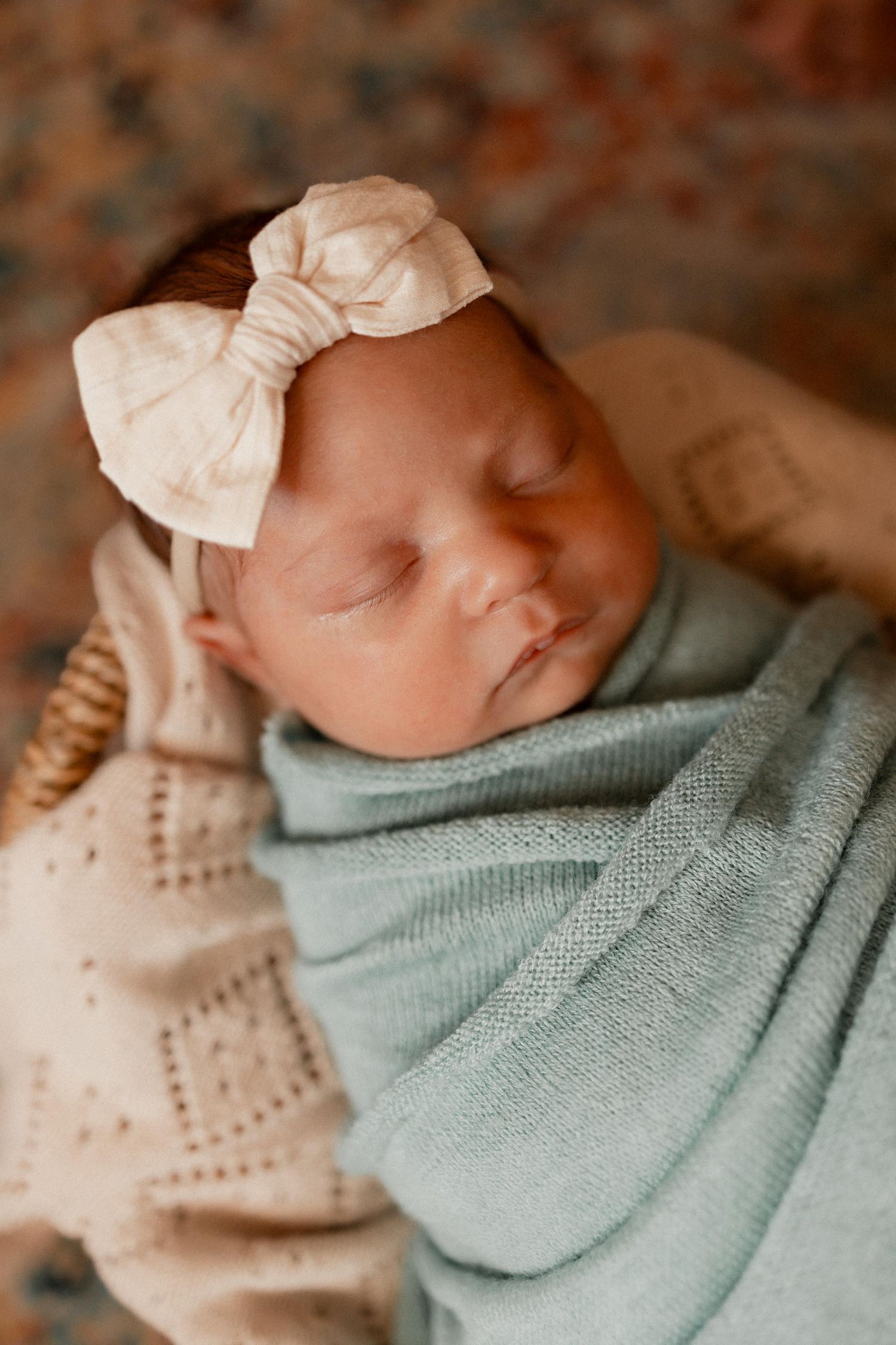 Peaceful newborn baby girl sleeping swaddled in mint green wrap with cream bow headband in wicker basket.