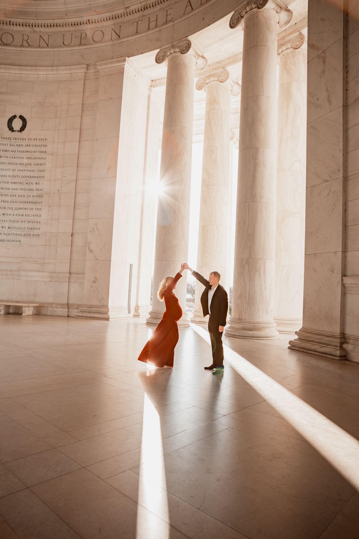 Two dancers perform in a sunlit classical building interior with dramatic shadows and columns.