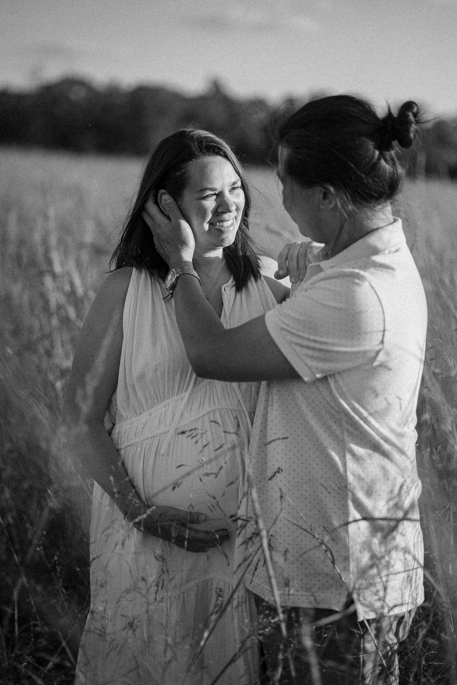 A couple in white clothing embraces and dances together in a sunlit meadow, captured in black and white photography.
