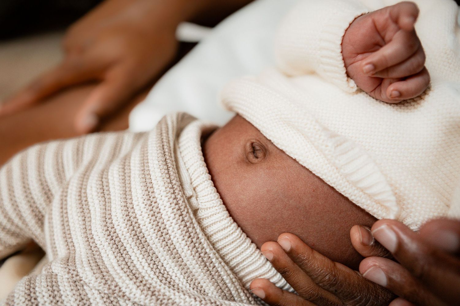 Newborn baby wearing white knit sweater and hat rests peacefully while being gently touched.
