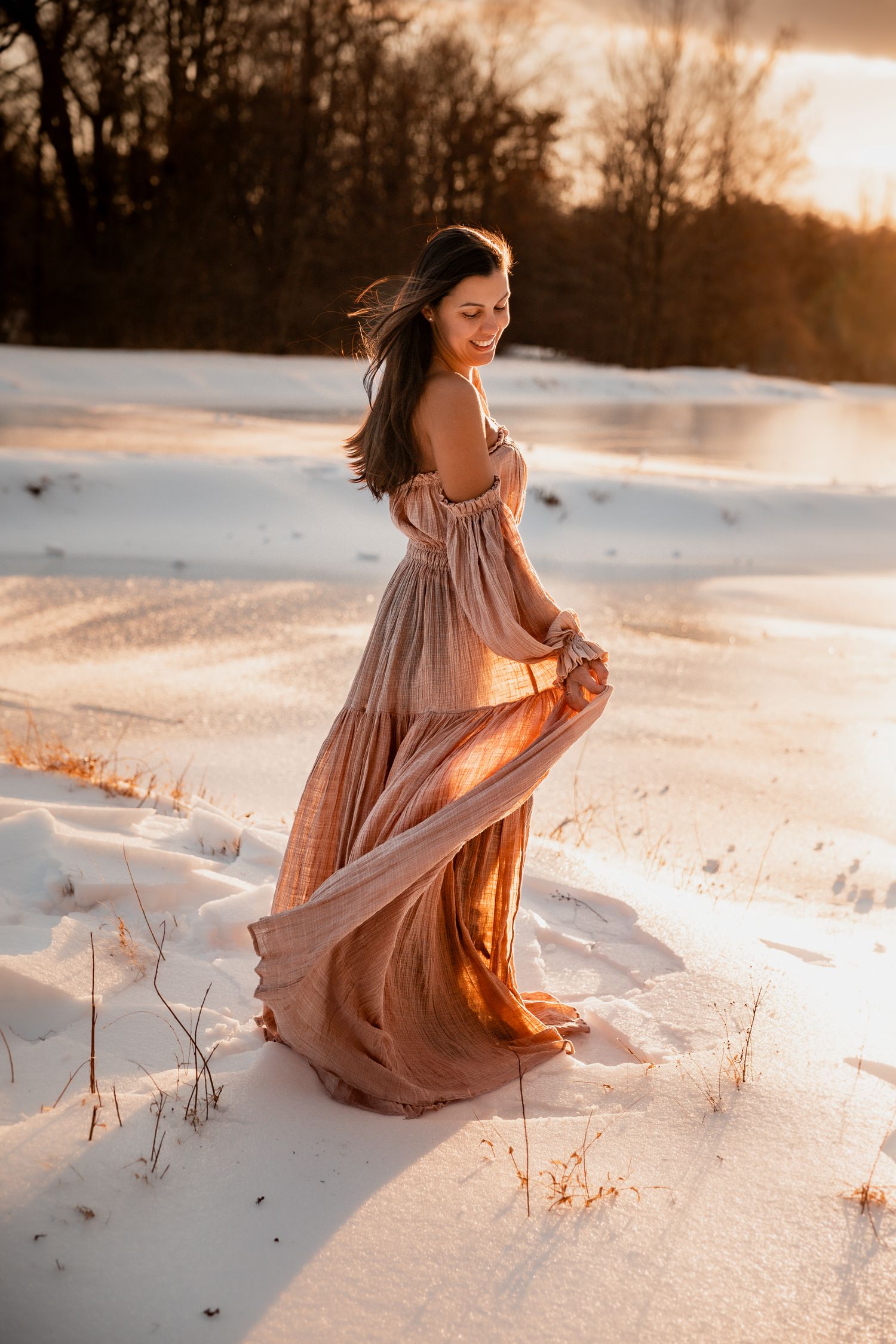 A person in a flowing peach dress sits gracefully in snowy field at sunset with warm golden light illuminating the scene.