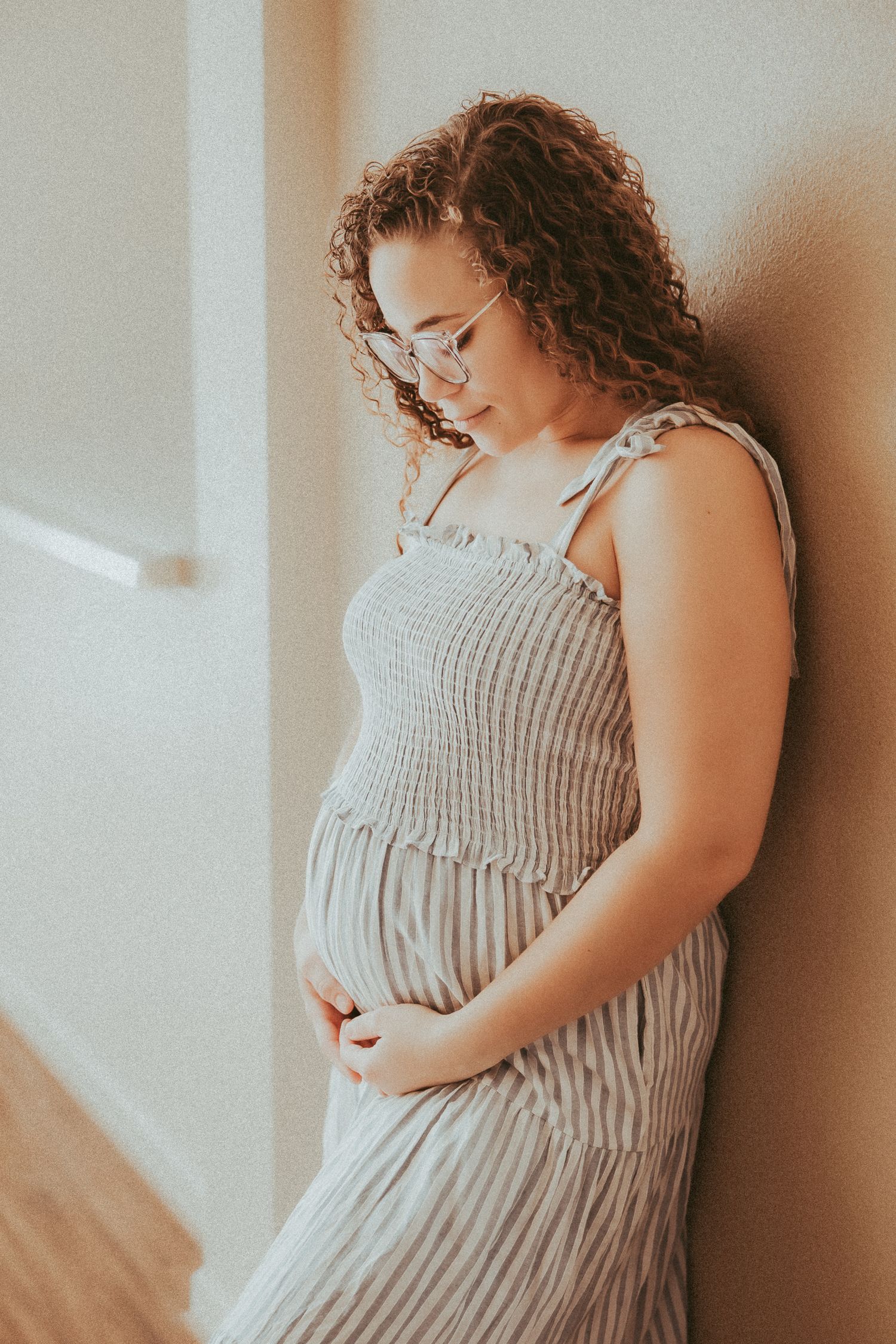 A pregnant woman in a soft gray dress stands by a window in warm natural light, gently cradling her baby bump.