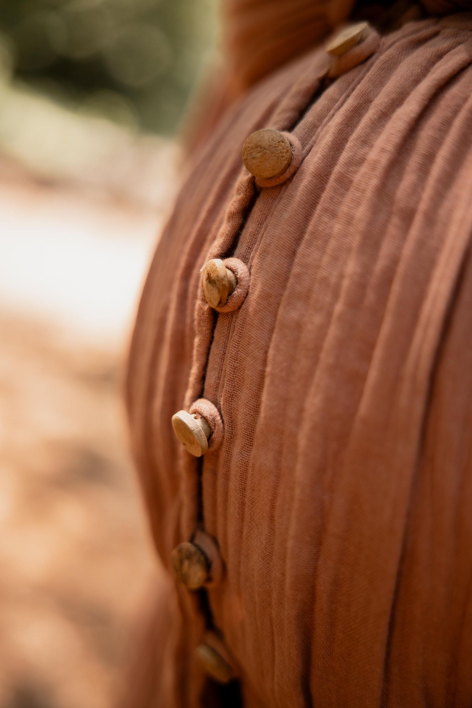 Close-up of rust-brown pleated fabric with wooden buttons on a leather cord, set against a blurred outdoor background.