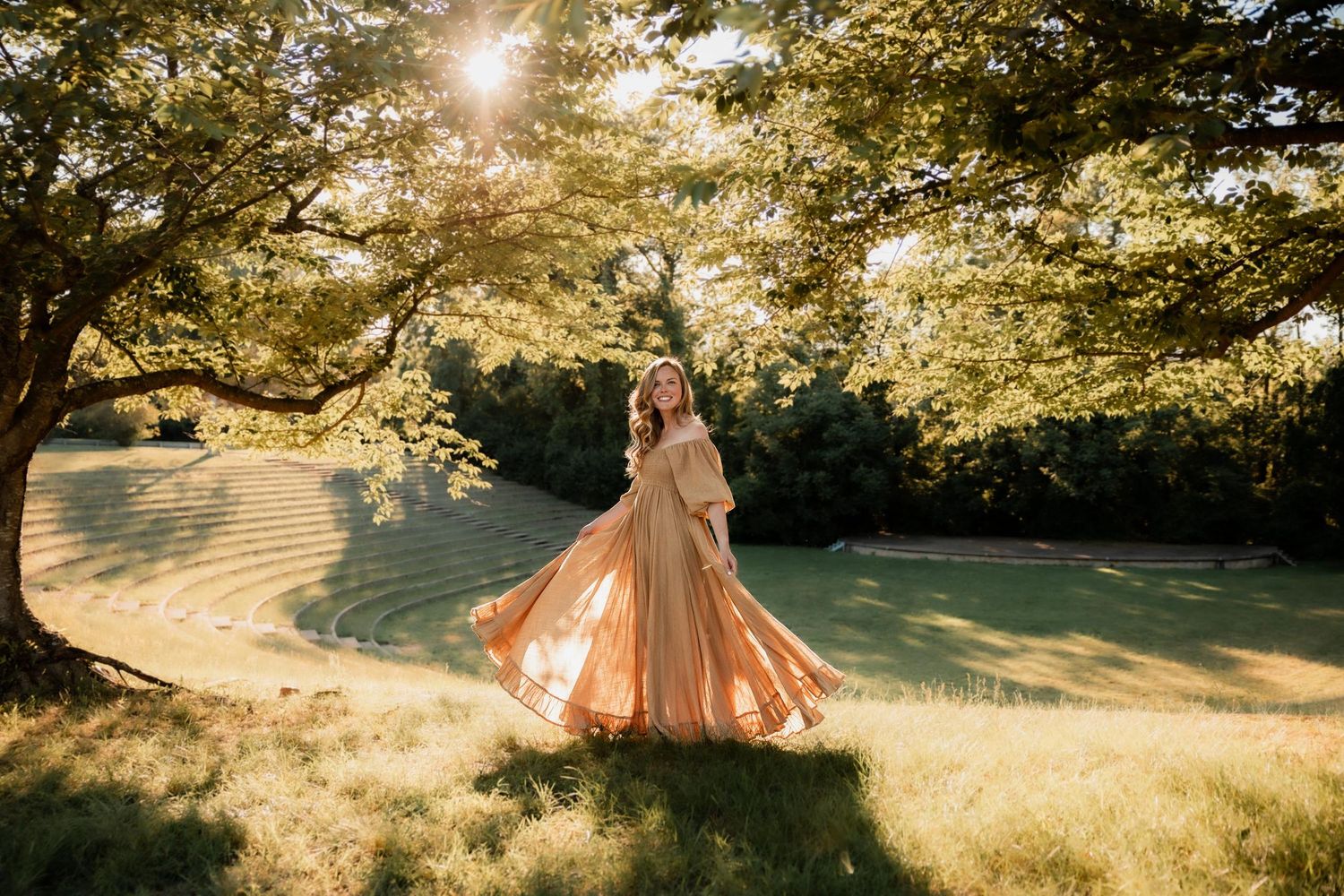 Woman in golden gown twirling under sunlit trees on lush green lawn.