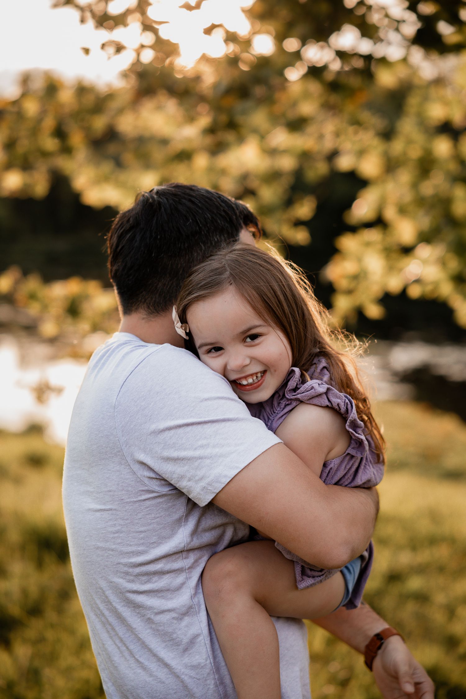 A smiling young girl hugs a man outdoors during golden hour, surrounded by lush green trees near a pond.