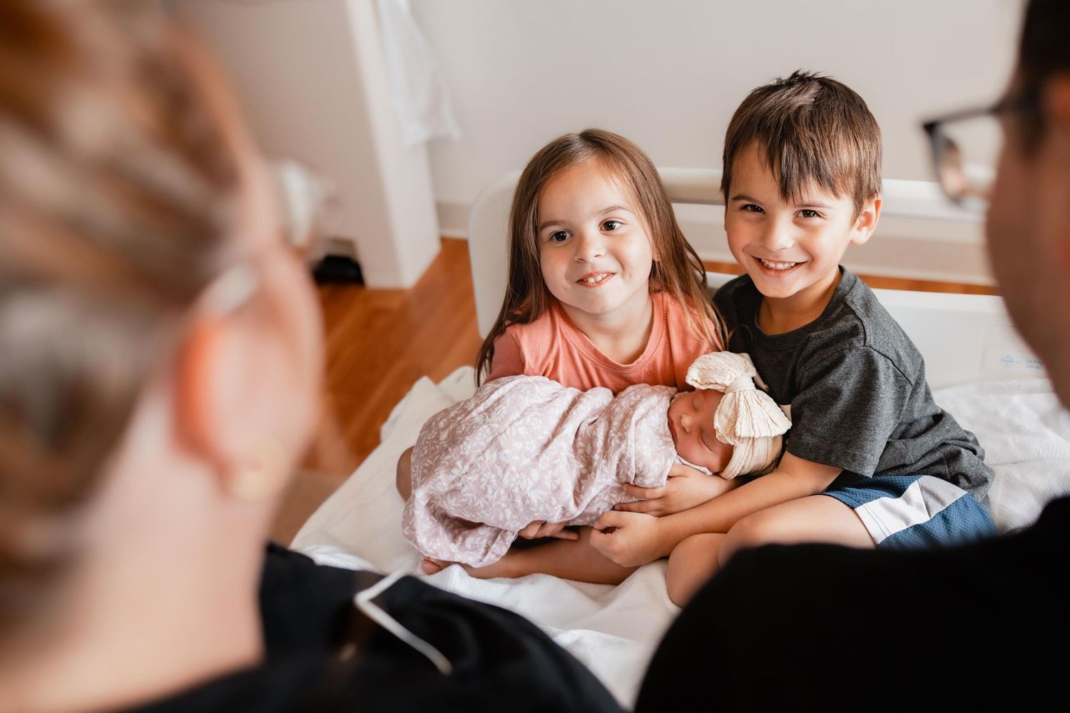 Two young siblings smiling and holding a newborn baby in a hospital room during a family visit.