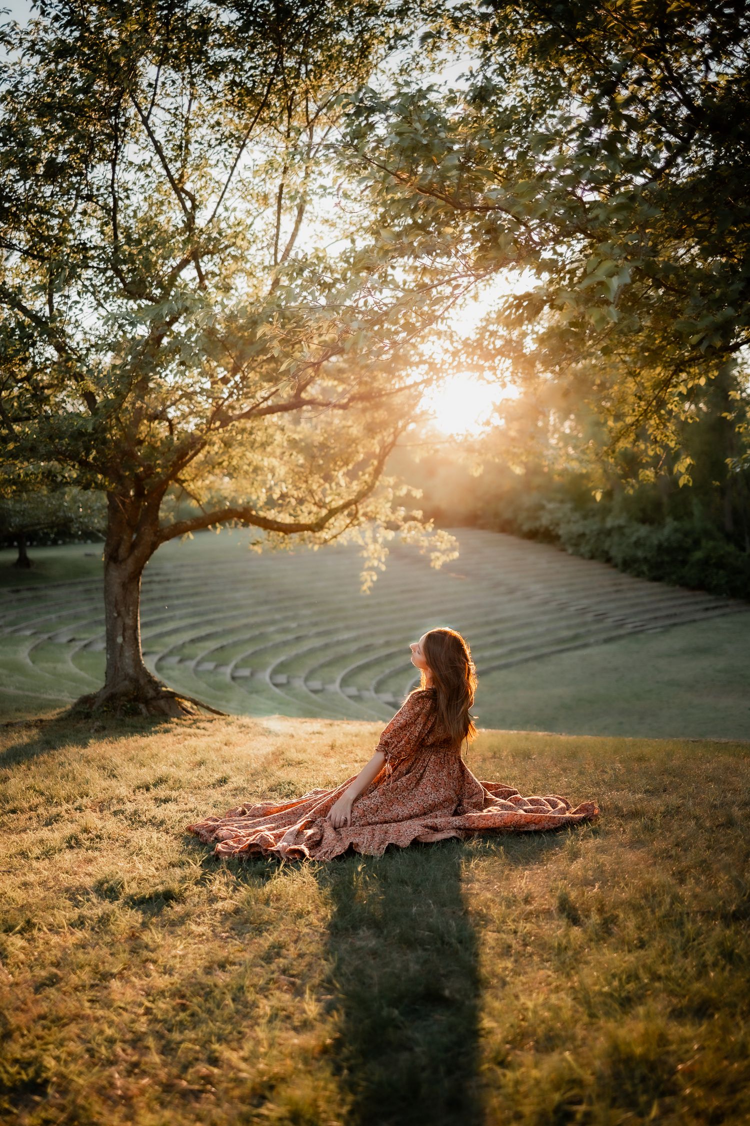 Woman in floral dress sitting on blanket under tree at golden sunset in lush green park.