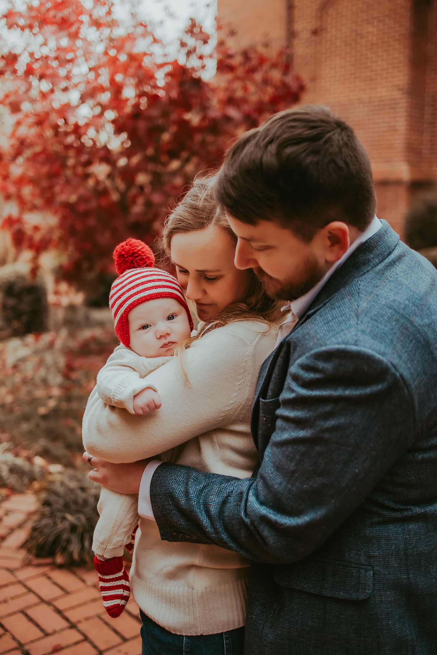 Family embraces in autumn setting with red foliage and brick backdrop.