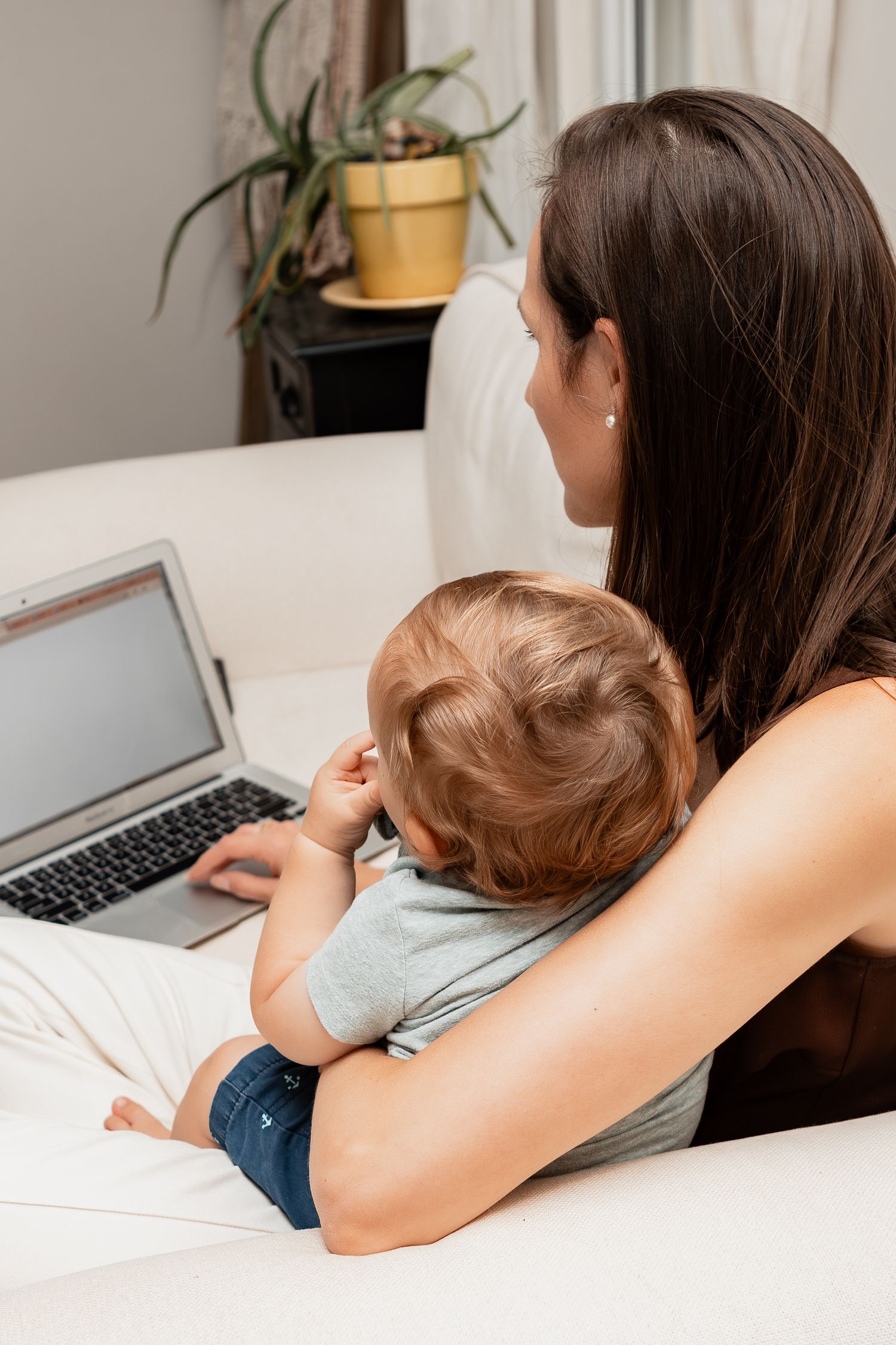 A parent and toddler looking at a laptop screen together while sitting on a white couch near a potted plant.