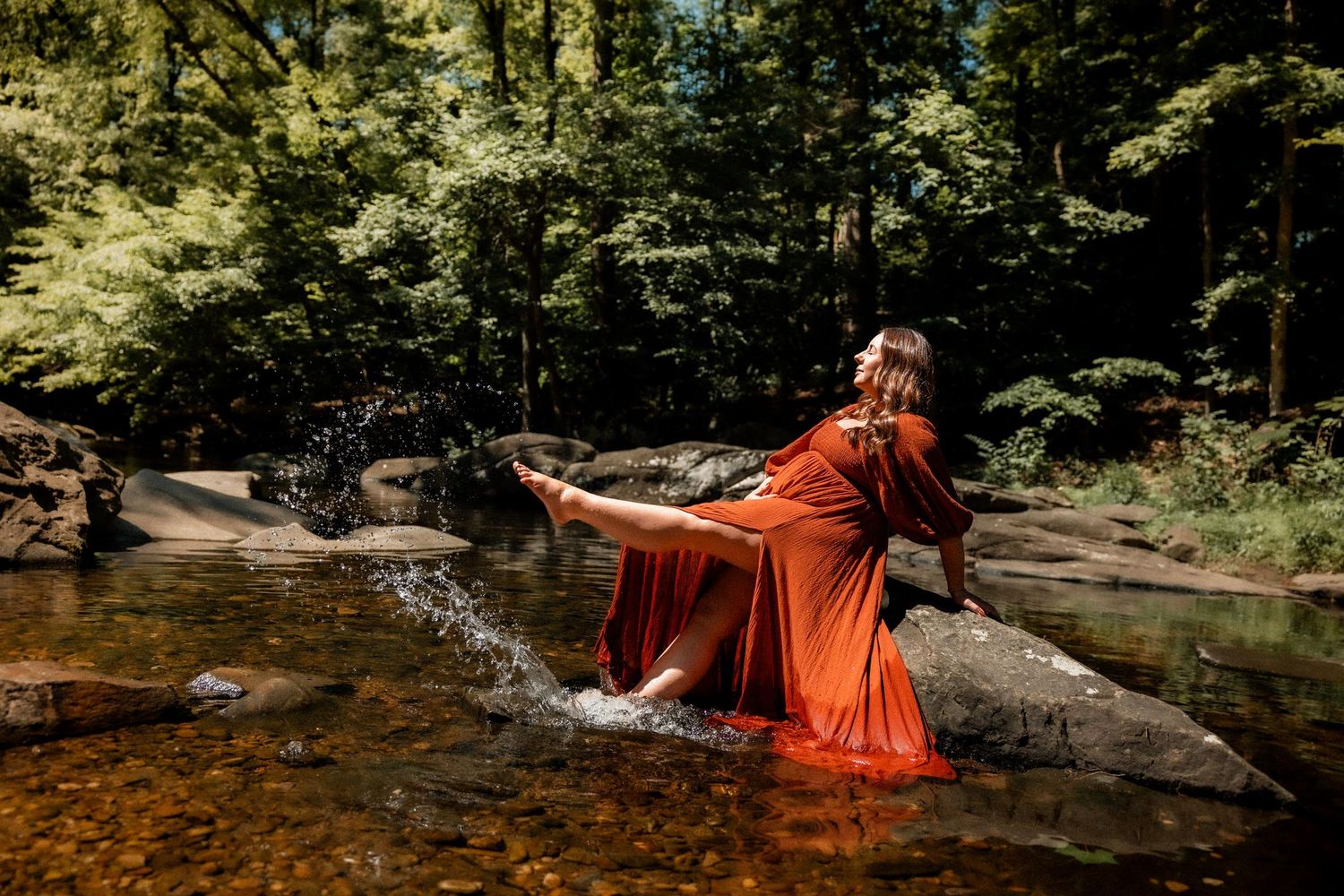 Woman in flowing rust-red dress splashing in a rocky forest stream surrounded by lush green trees.