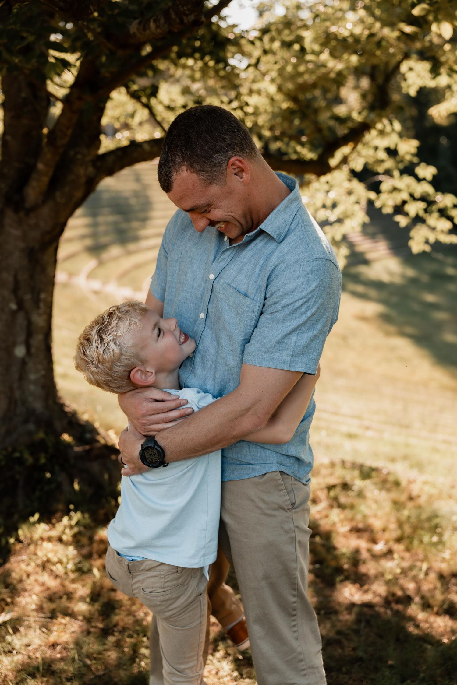 A father and young blonde son share a warm embrace outdoors under a tree in golden sunlight.