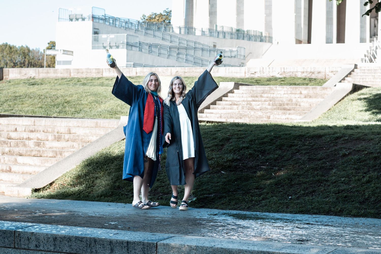 Two graduates in blue caps and gowns celebrate with raised arms at memorial steps.