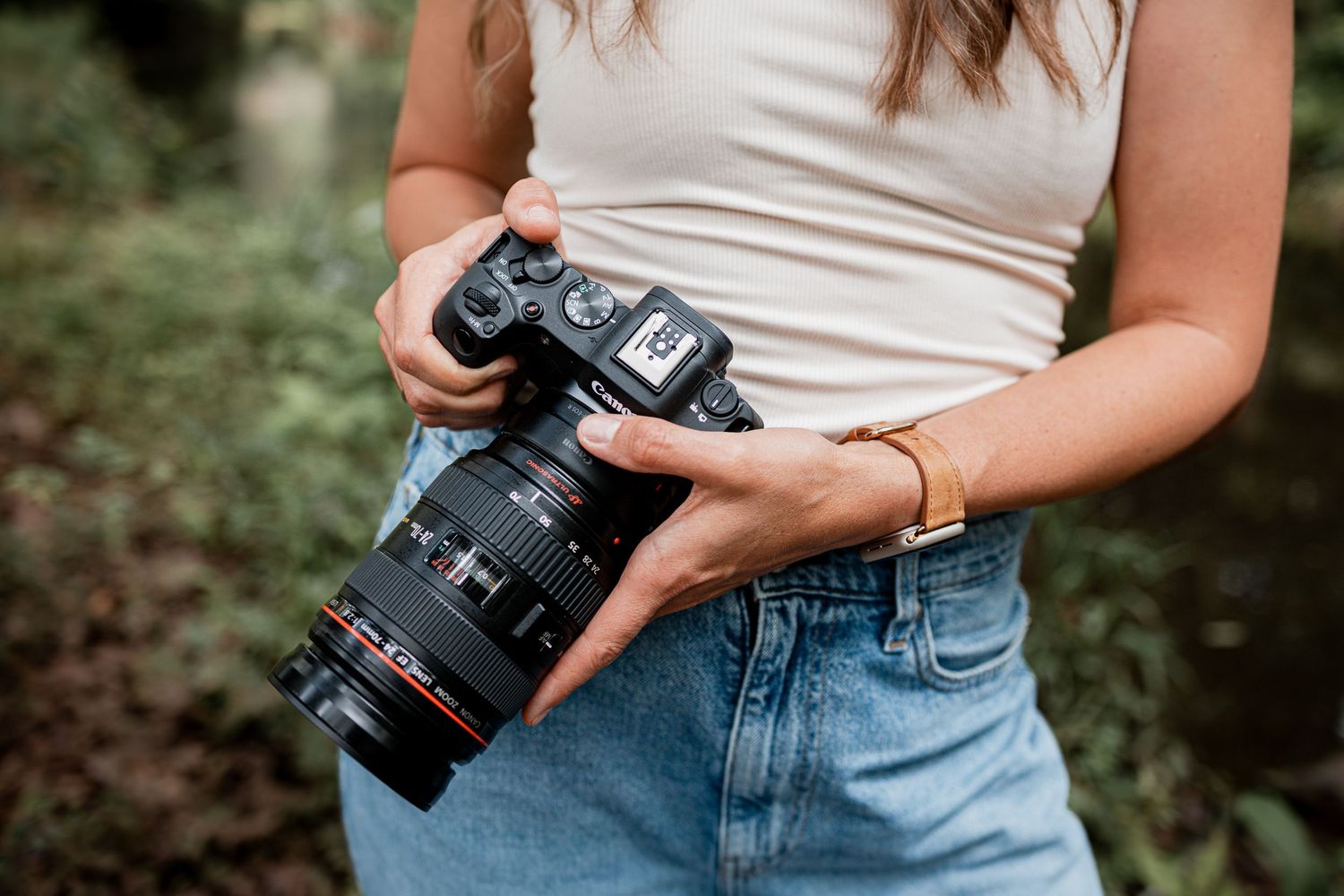 Person in blue jeans and white top holding professional camera outdoors in natural setting.