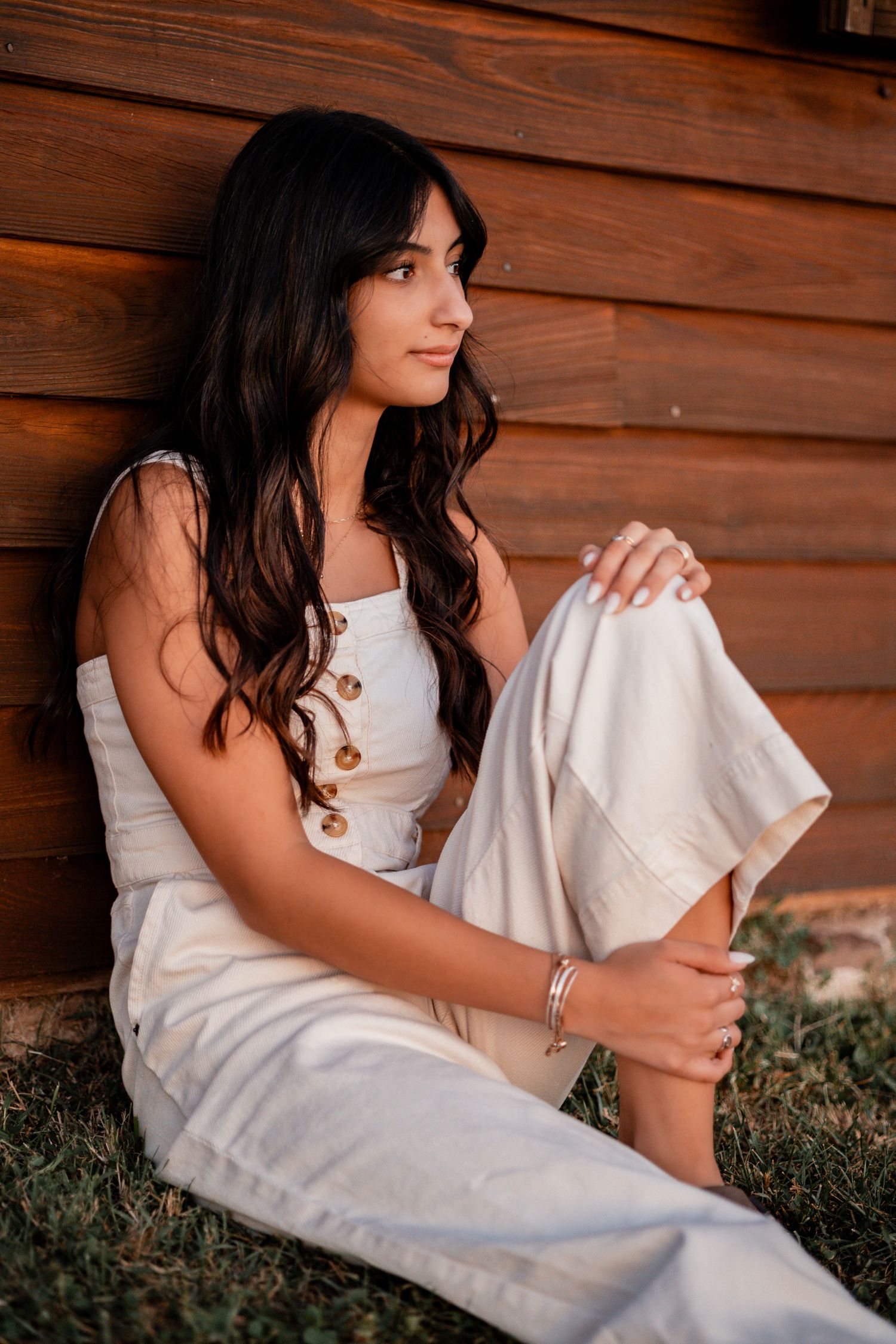 A woman in white clothing sits beside a rustic wooden wall during sunset.