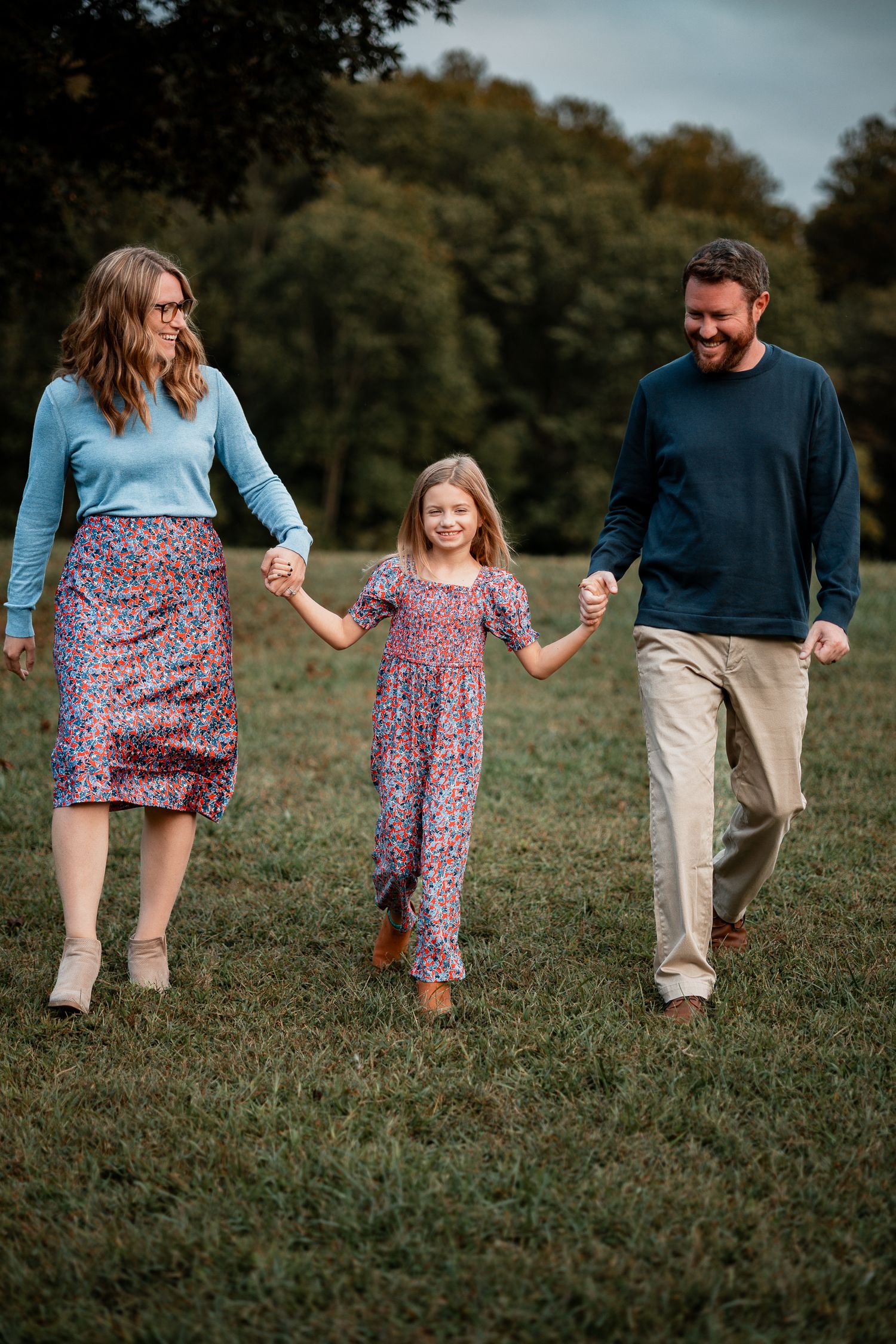 Family wearing coordinating floral outfits walks together holding hands in a grassy field on an autumn day.