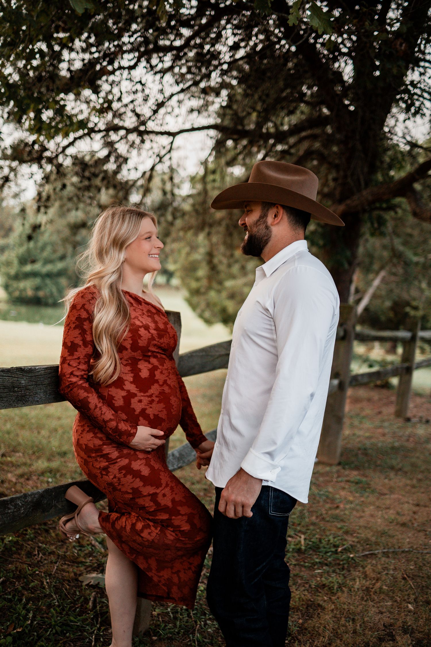 A couple in a rustic setting, with her wearing a red dress and him in a cowboy hat leaning against a wooden fence.