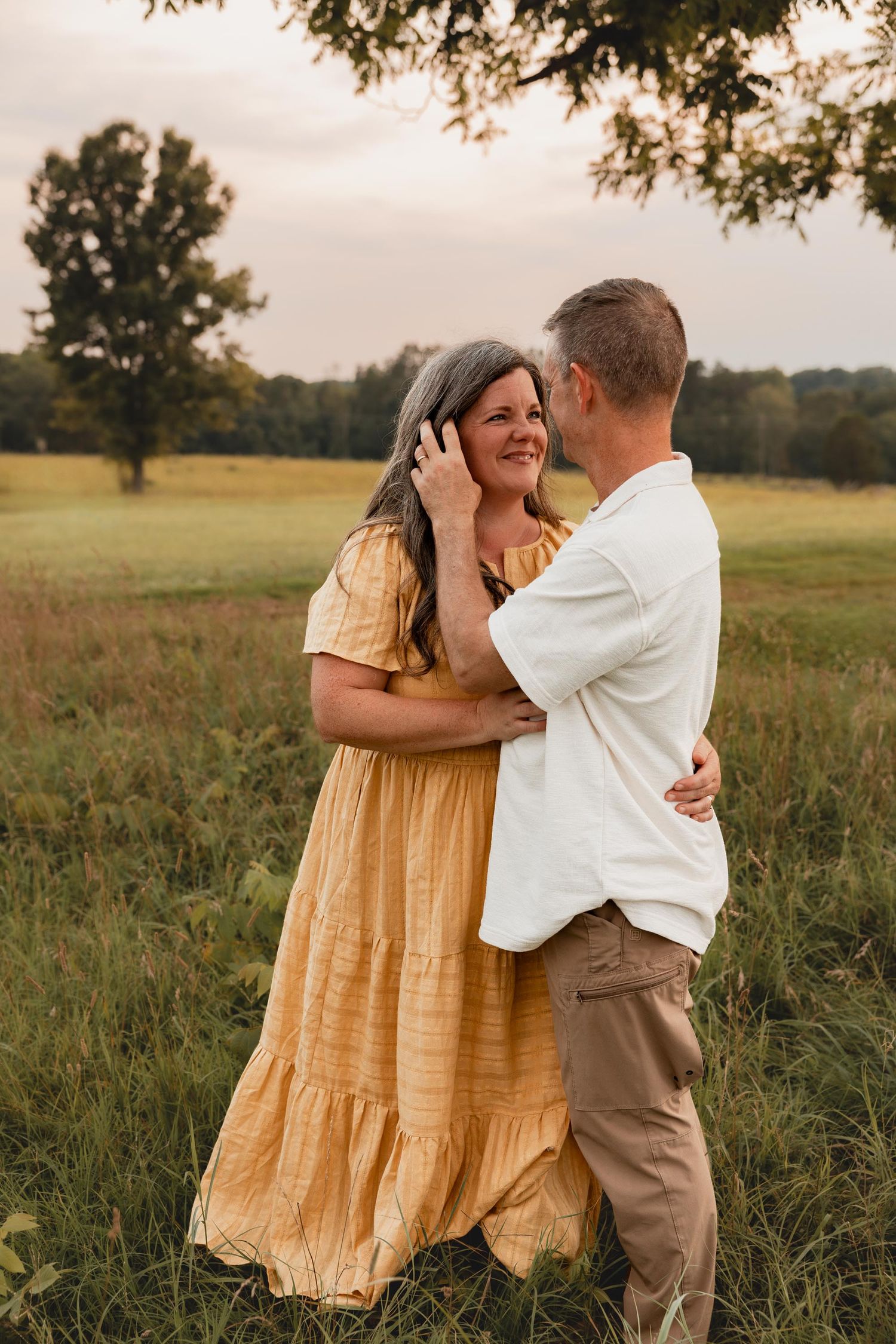 Couple embracing in a golden field, woman in yellow dress and man in white shirt, sharing a romantic moment outdoors.