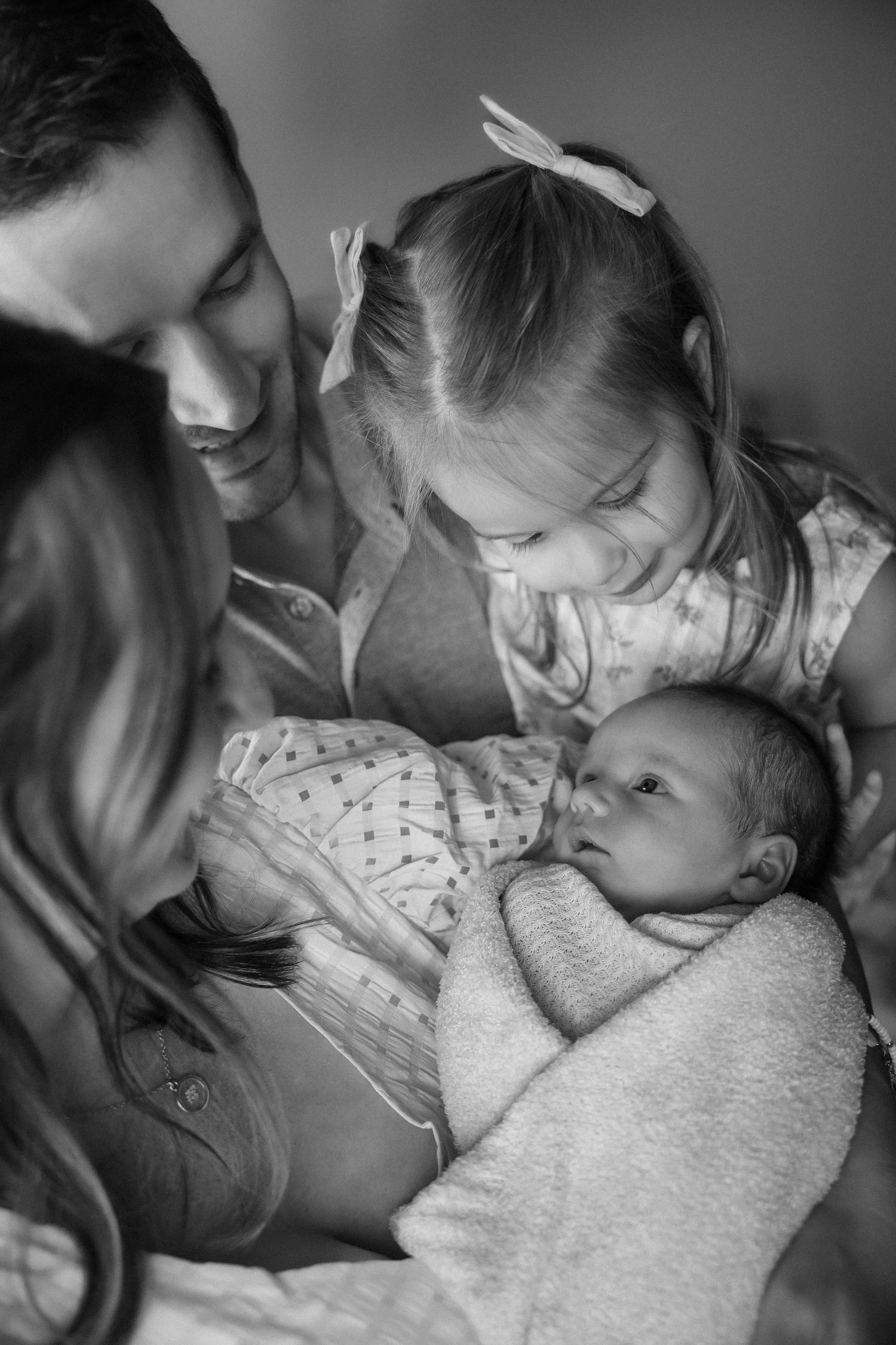 Black and white family photo of parents and young daughter smiling and admiring their swaddled newborn baby.