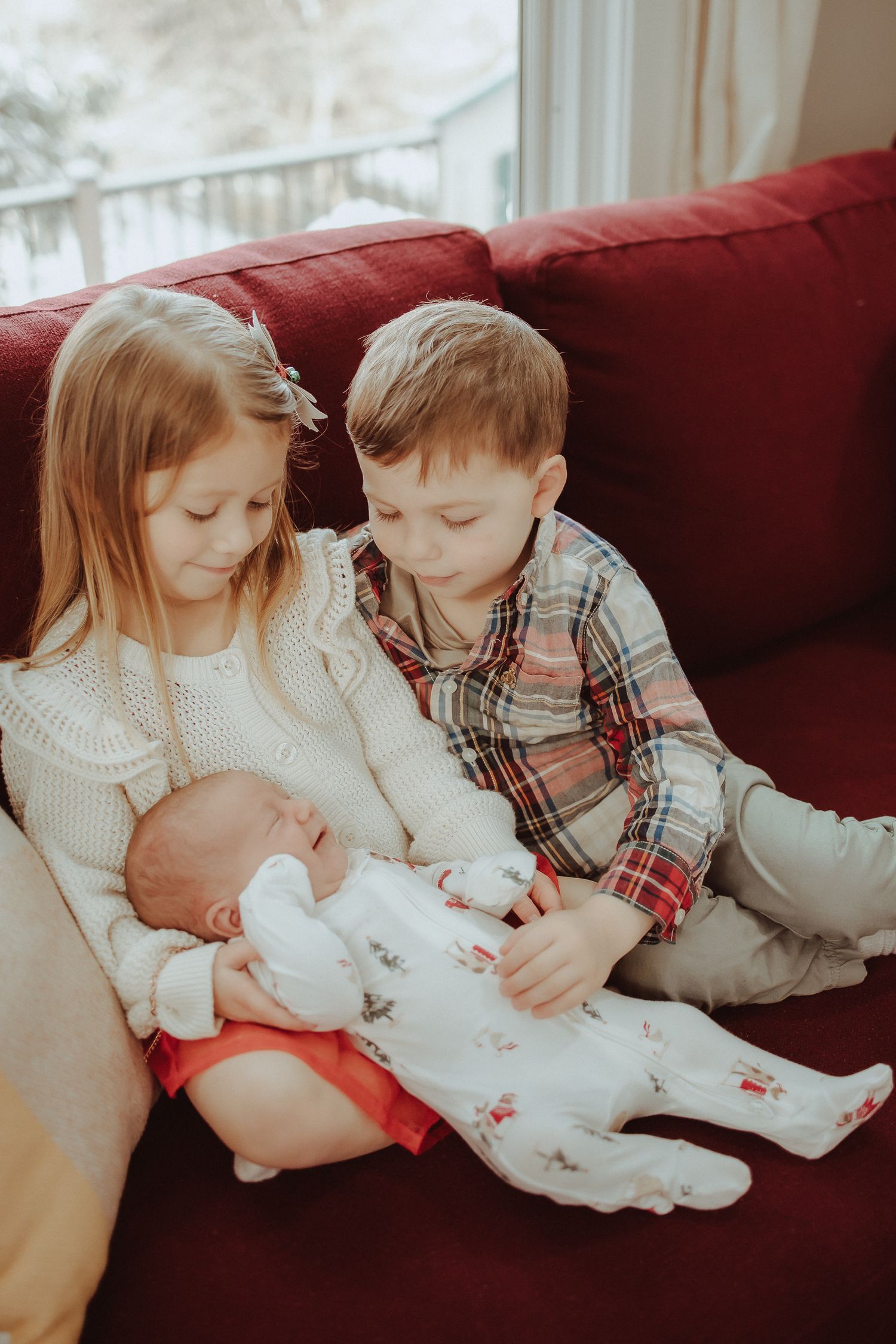 Two young children in winter attire lovingly embrace and interact with a newborn baby on a red couch.