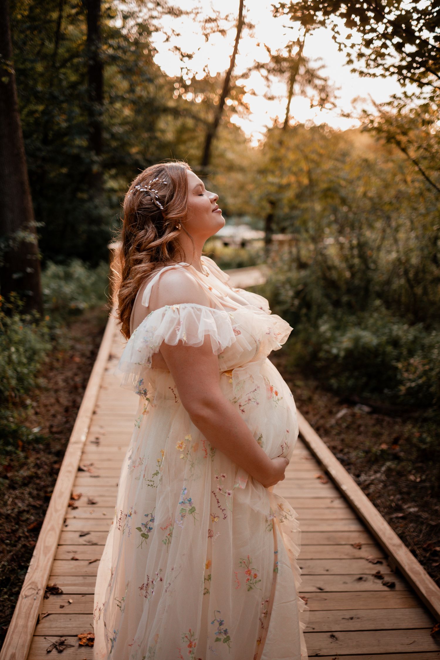 A figure in a flowing white floral dress stands on a wooden boardwalk path during golden hour in a forest setting.