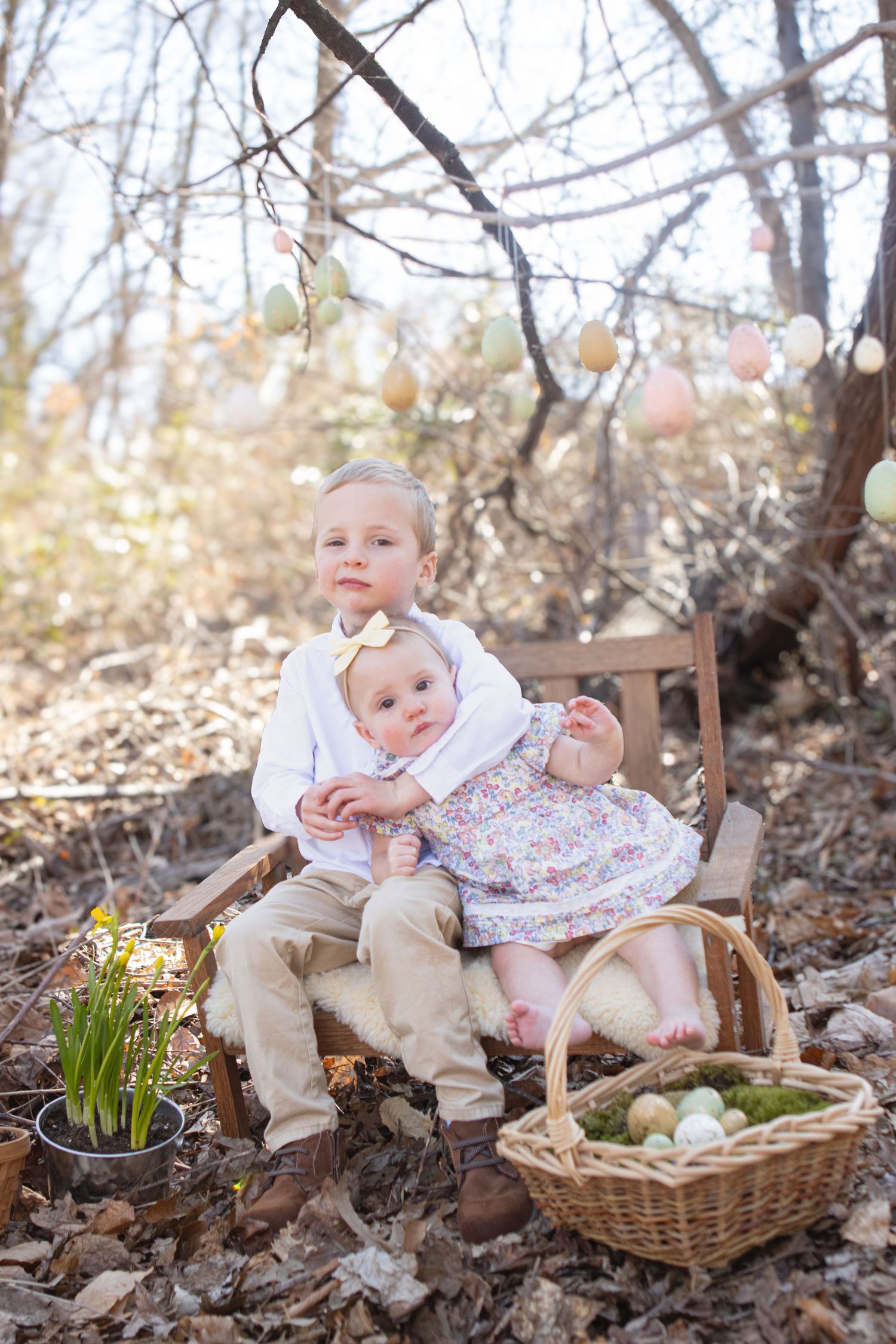 Two children sit on a wooden chair outdoors with Easter decorations and spring flowers nearby.