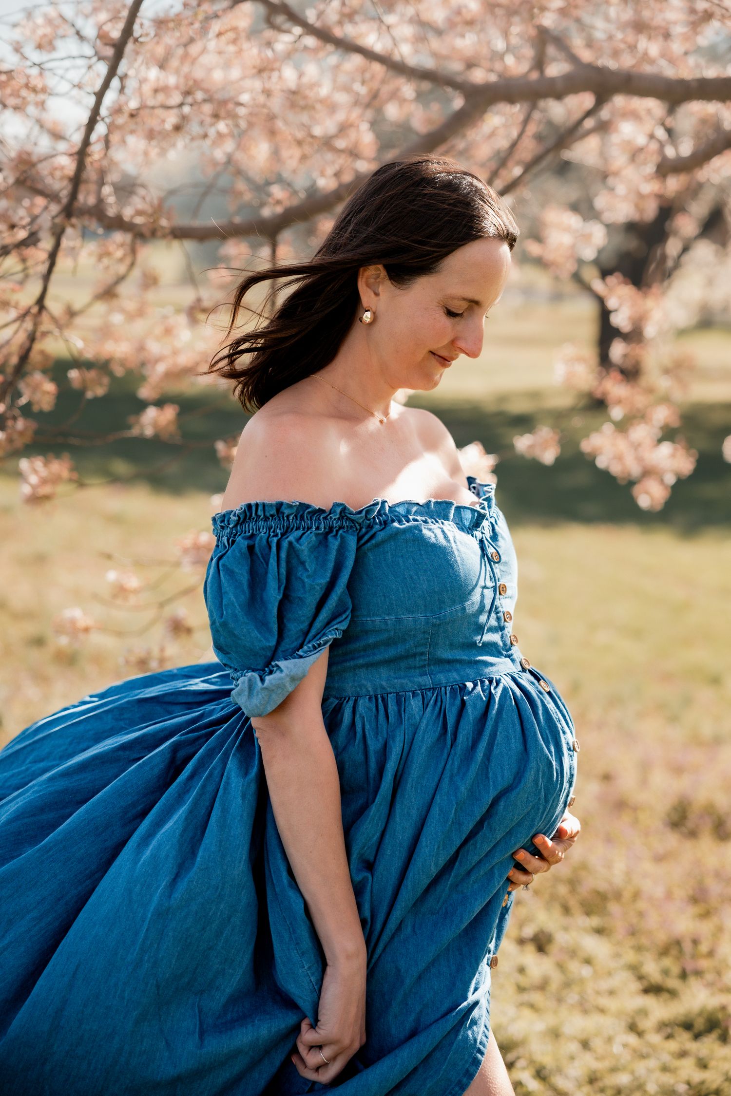 Pregnant woman in blue off-shoulder dress holding baby bump near cherry blossom tree outdoors.