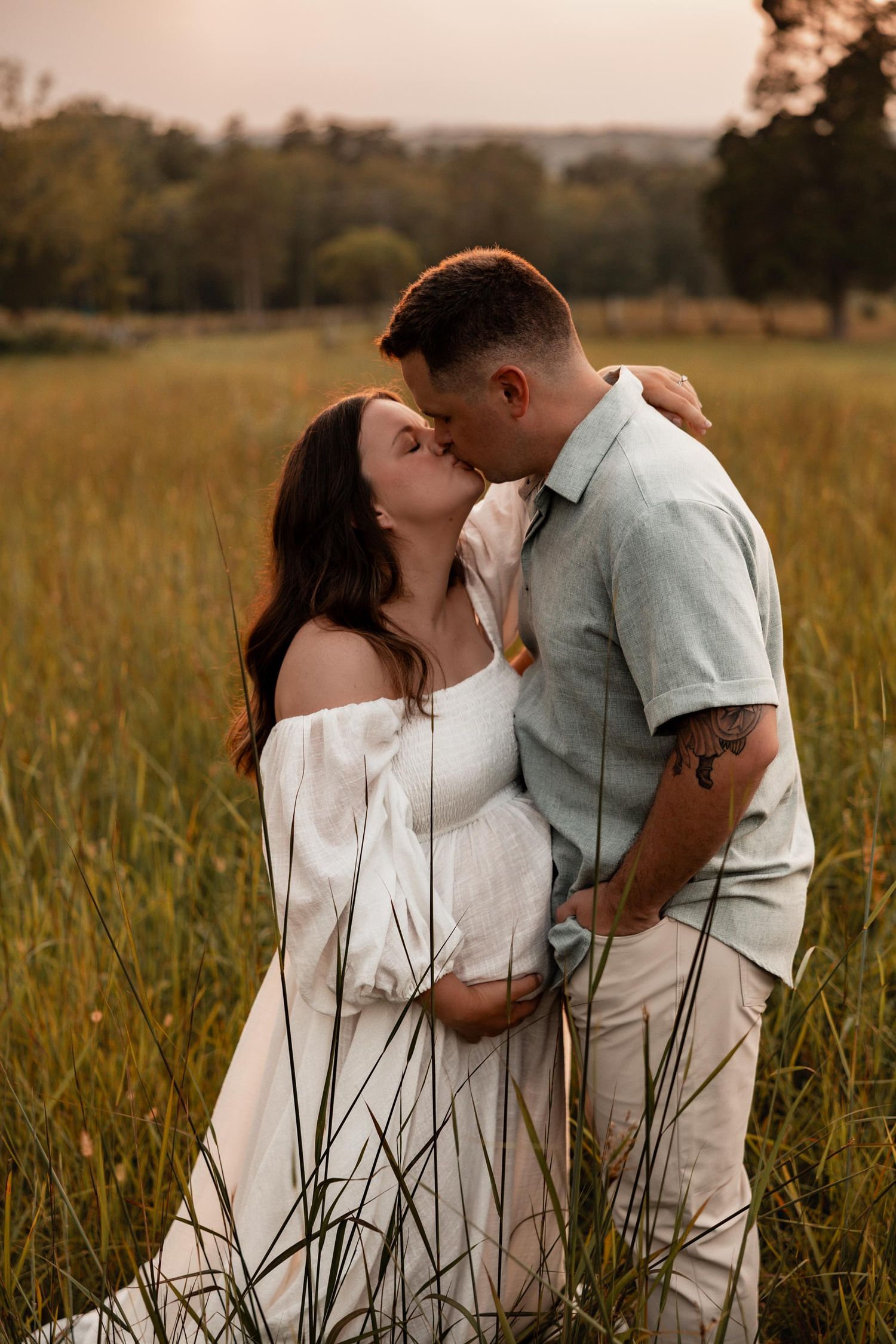 Expecting couple sharing a kiss in tall golden grass at sunset, man with tattoo embracing woman in white dress.