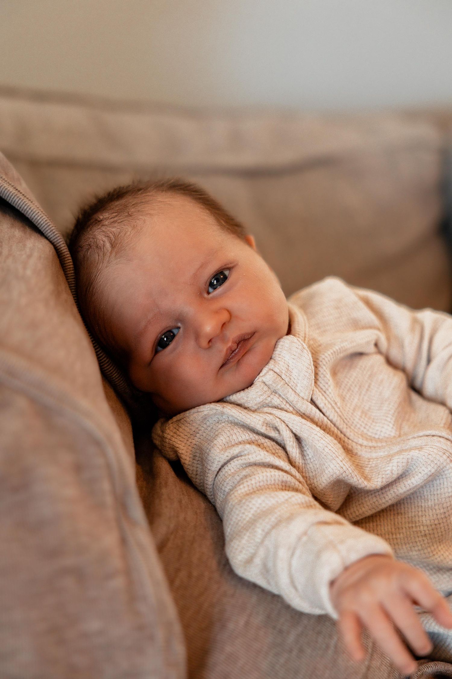 Newborn baby in a cream waffle onesie lying on a beige sofa, looking at the camera.
