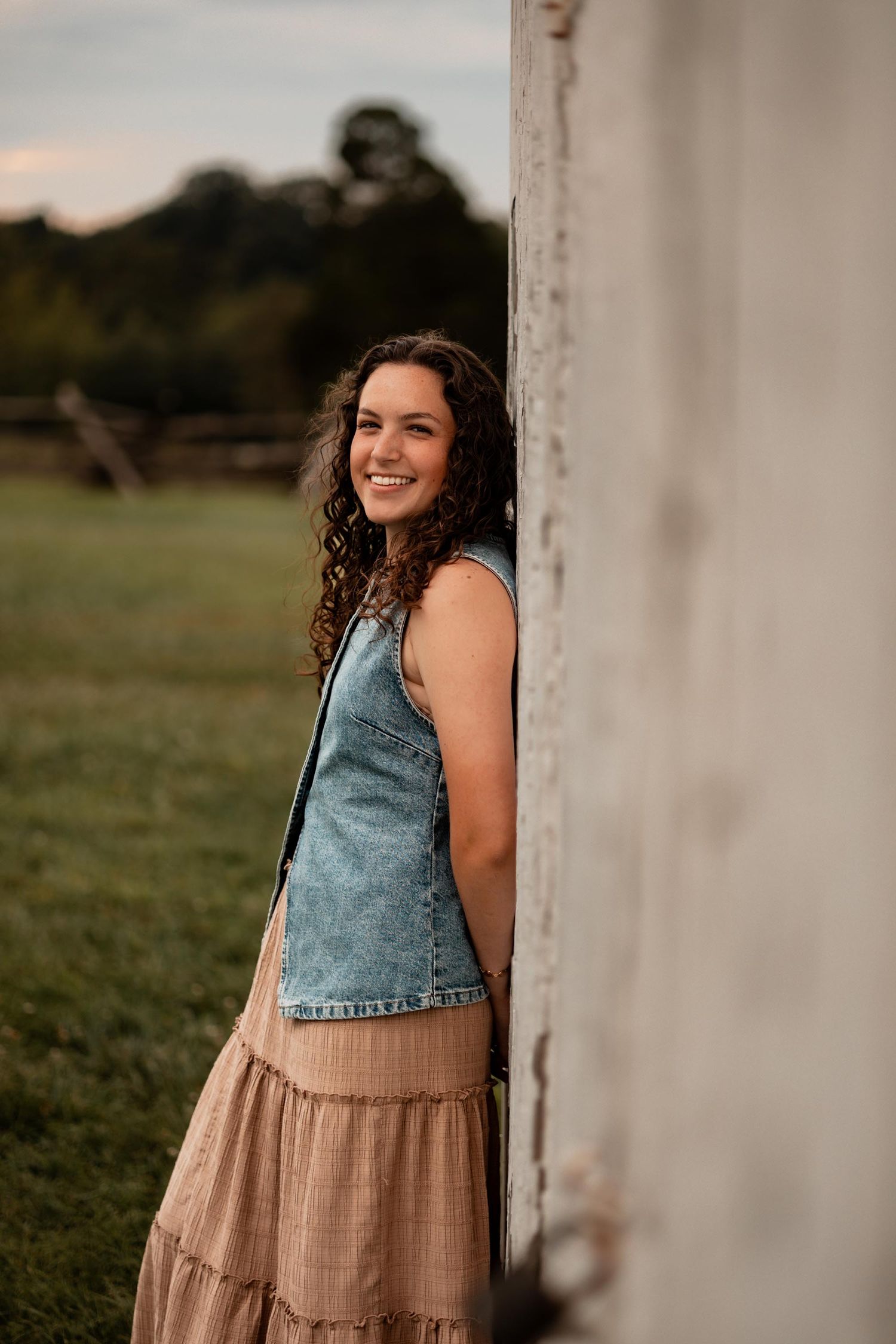 Young woman in denim vest and tan skirt leaning against white barn wall, smiling outdoors.