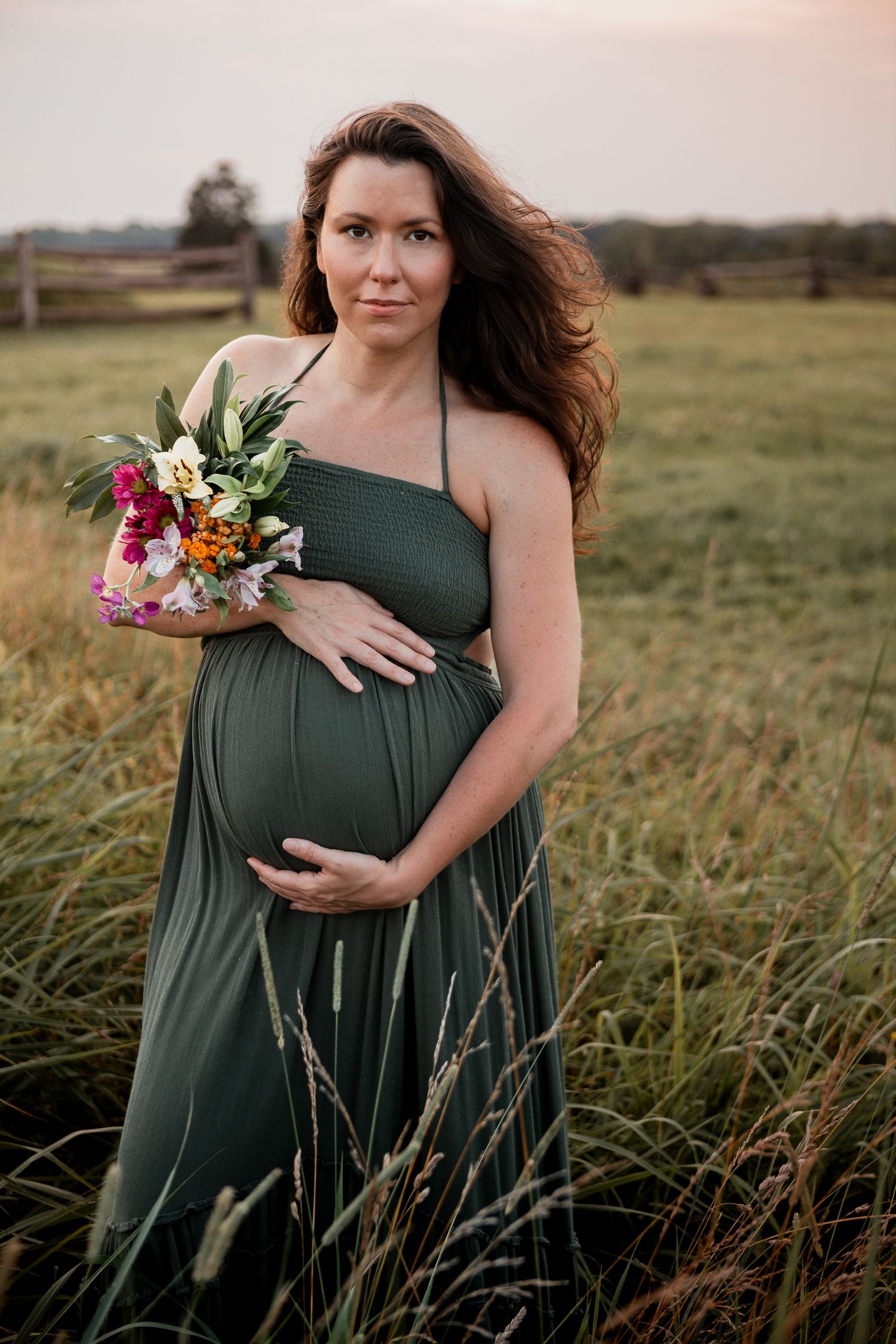 An expectant mother poses in a grassy field wearing a dark green halter dress while holding a colorful bouquet.