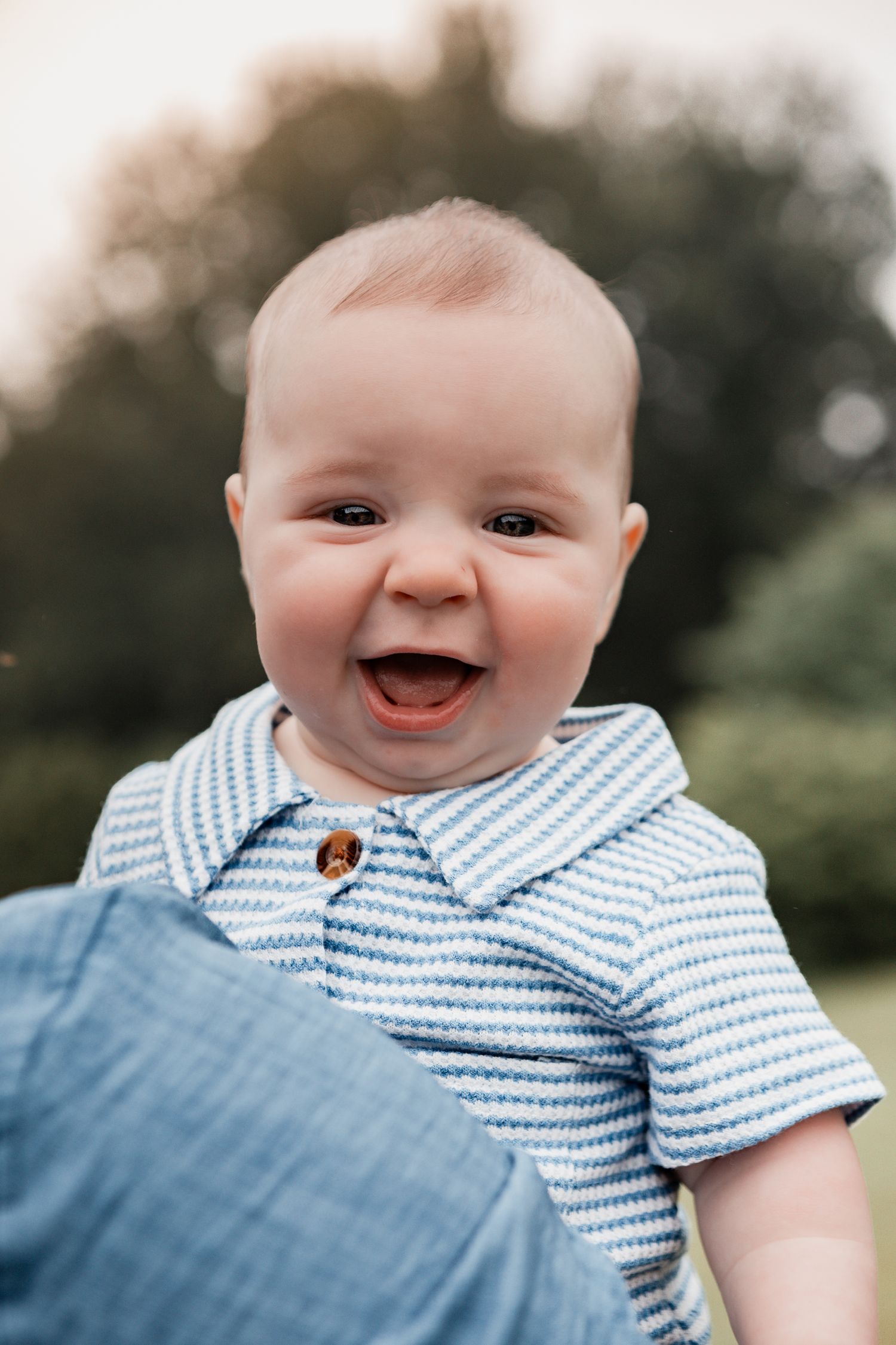 A happy baby in a blue striped collared shirt smiles broadly while sitting outdoors.