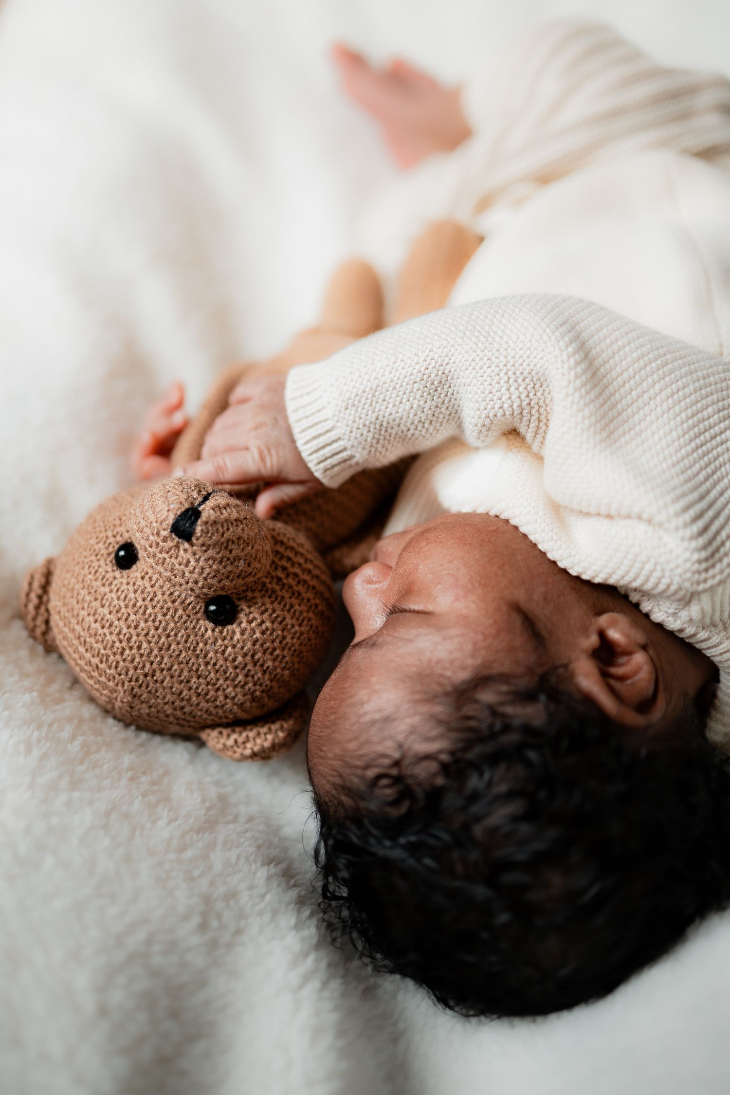 A sleeping infant lies peacefully next to a brown teddy bear under a white knit blanket.