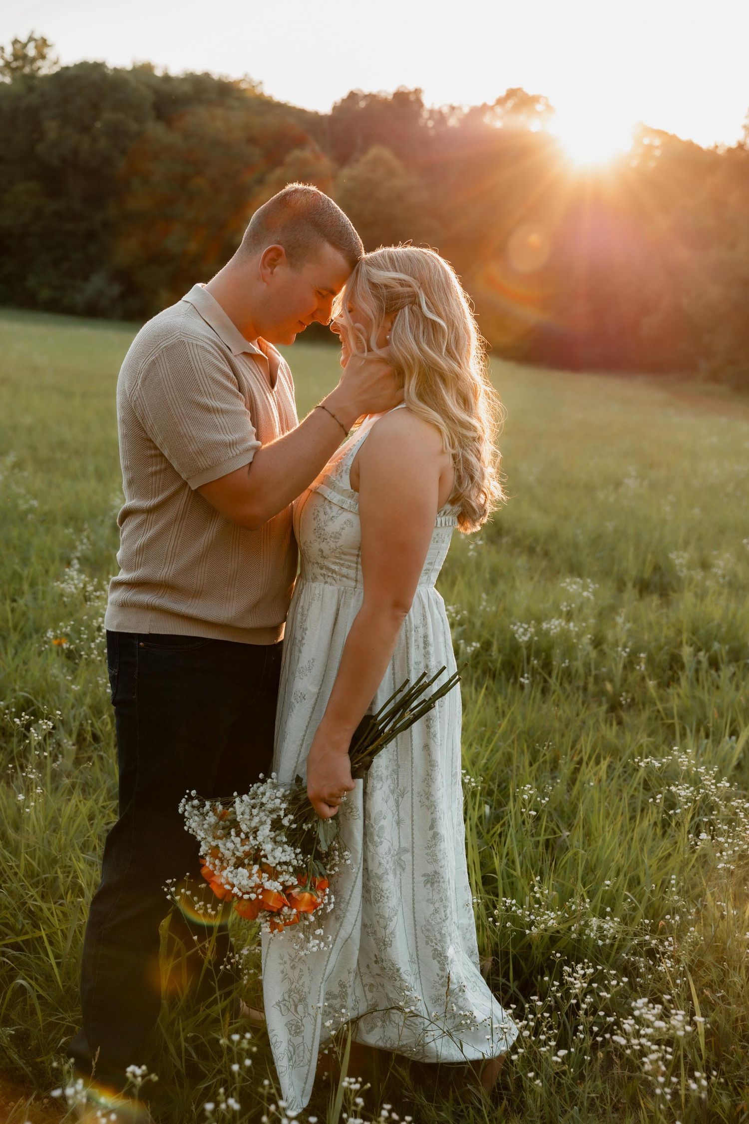Couple sharing a romantic moment in a sunlit green field, woman holding wildflower bouquet at golden hour.