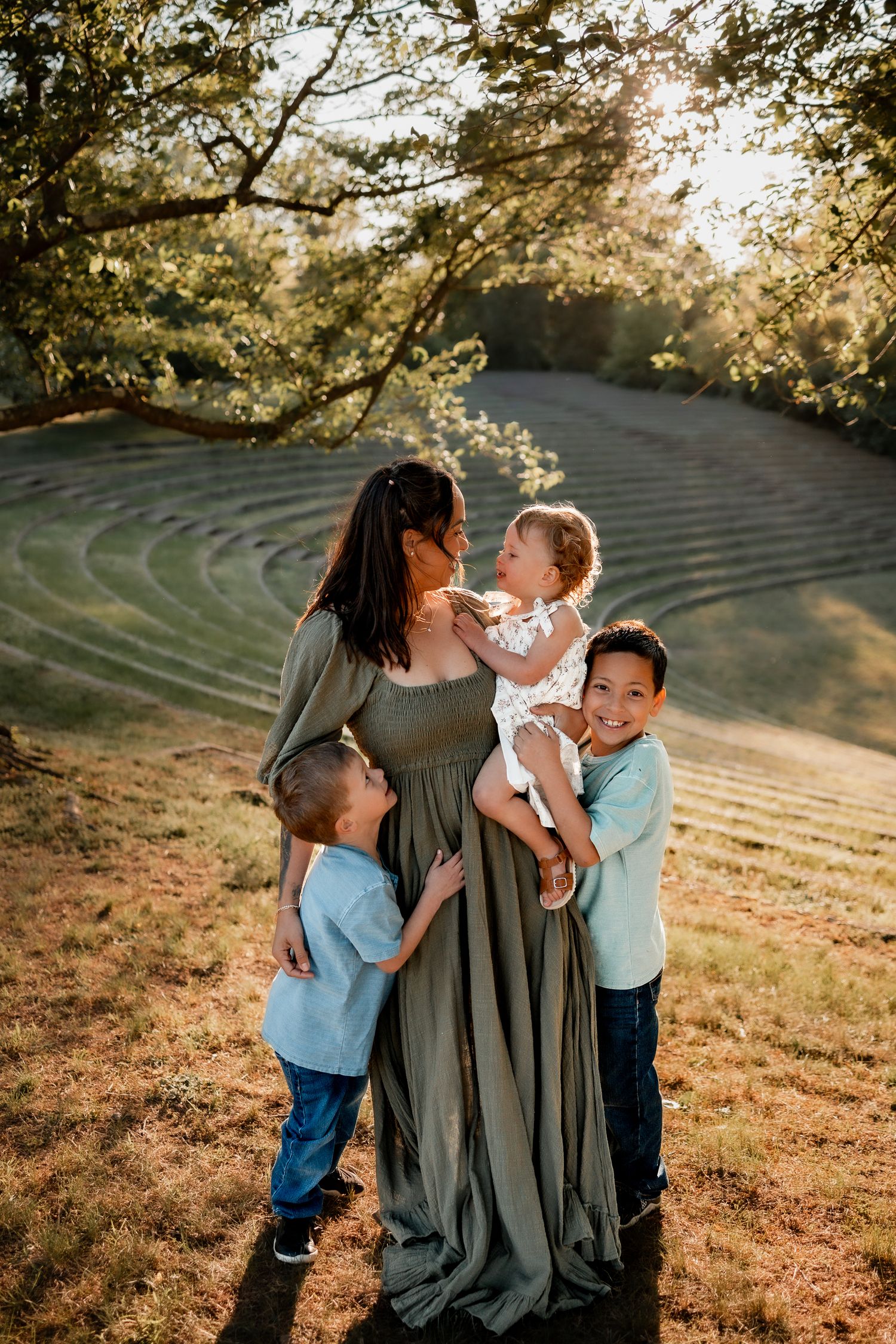 A mother in a green dress holds her baby while two young boys smile and hug her outdoors in golden sunlight.
