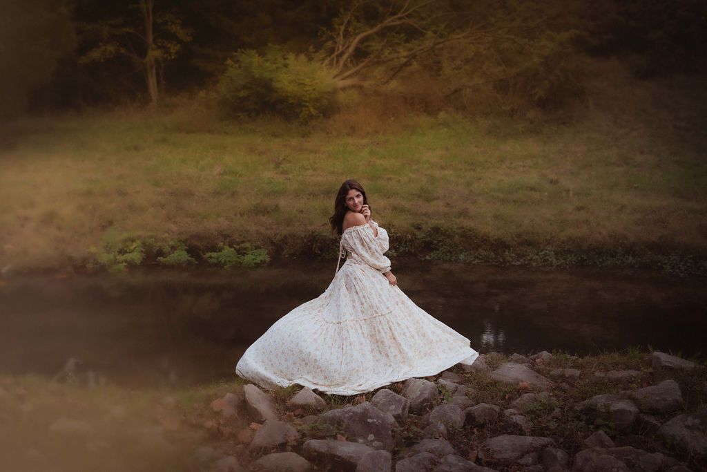 A woman in a flowing white lace gown poses gracefully on rocks near a serene forest stream at dusk.