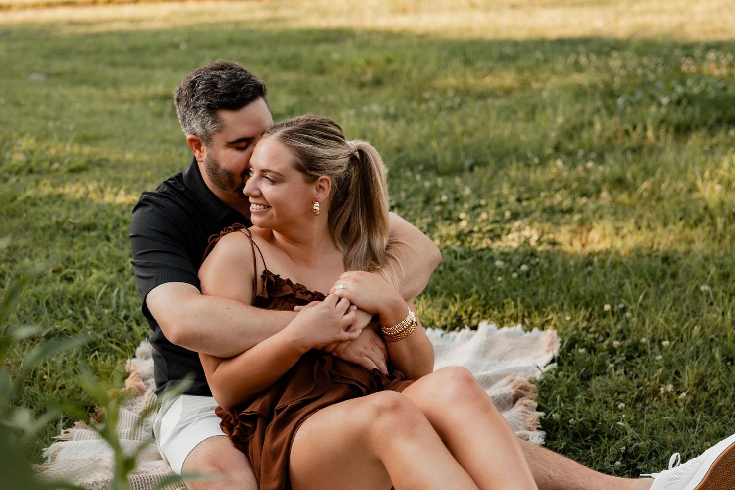 Couple sitting on a blanket in a green park, man embracing woman from behind, sharing a tender moment outdoors.