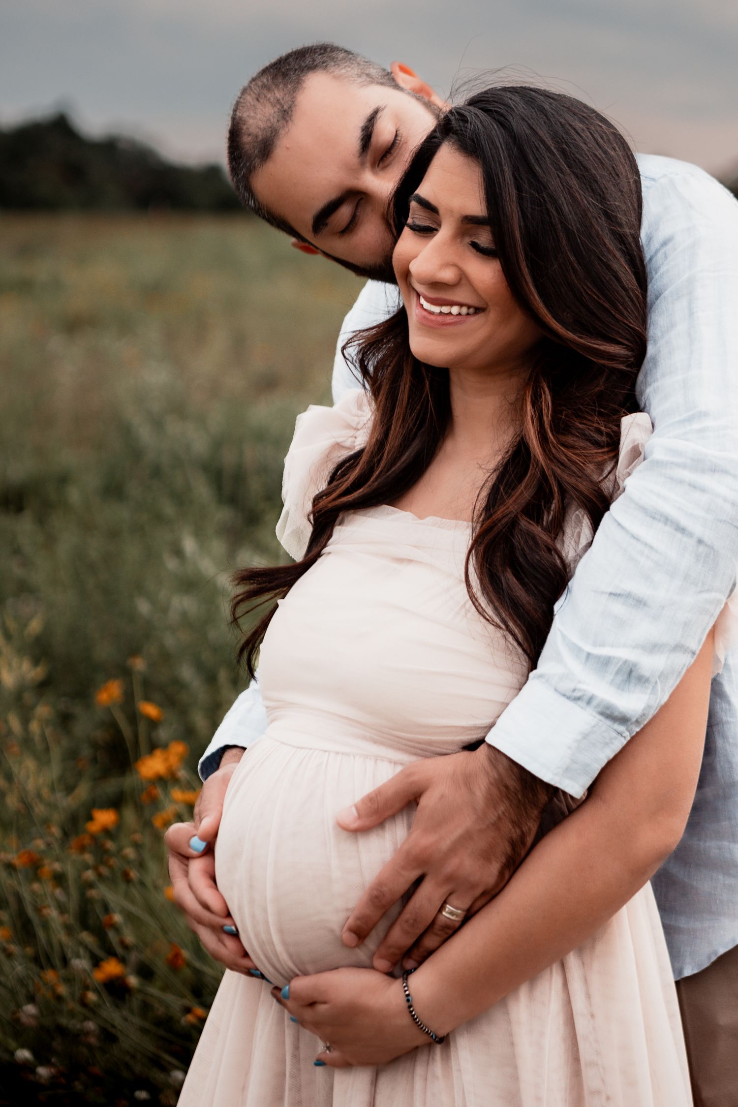 A loving maternity photo shows a couple embracing in a field of wildflowers at sunset.