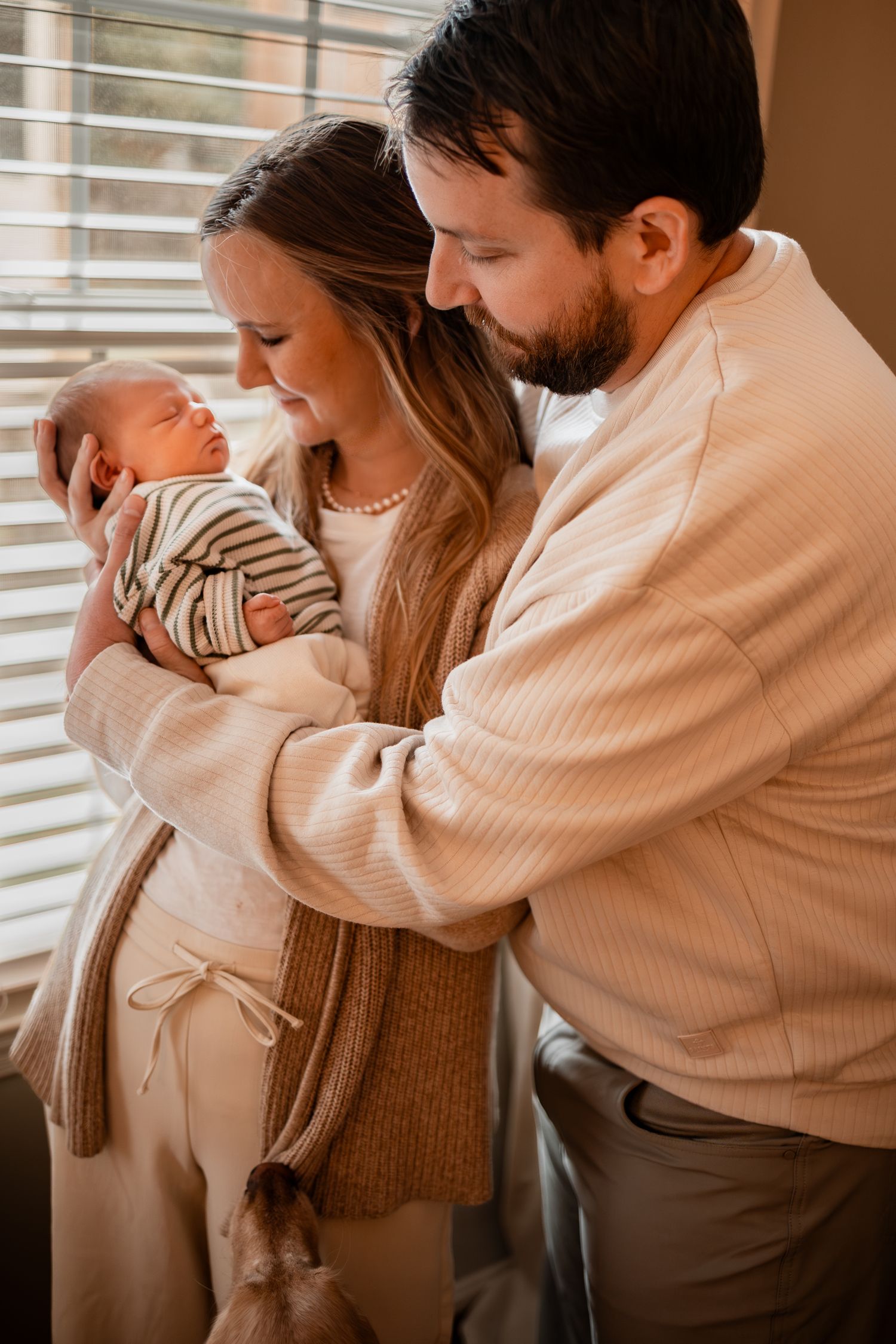 Couple in cozy sweaters holding their newborn baby near window.