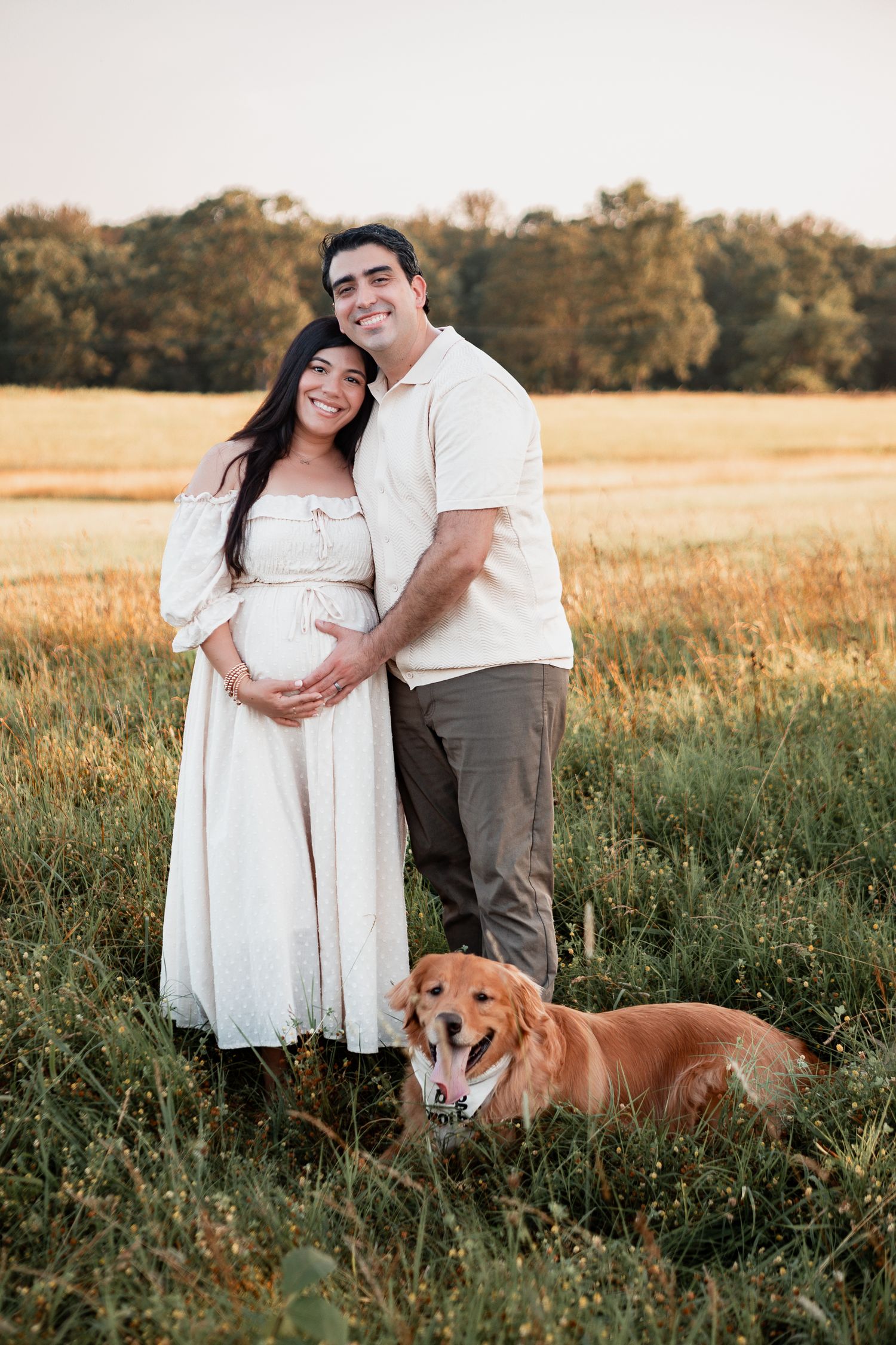 A couple in white clothing embraces in a golden field at sunset with their brown dog lying nearby.