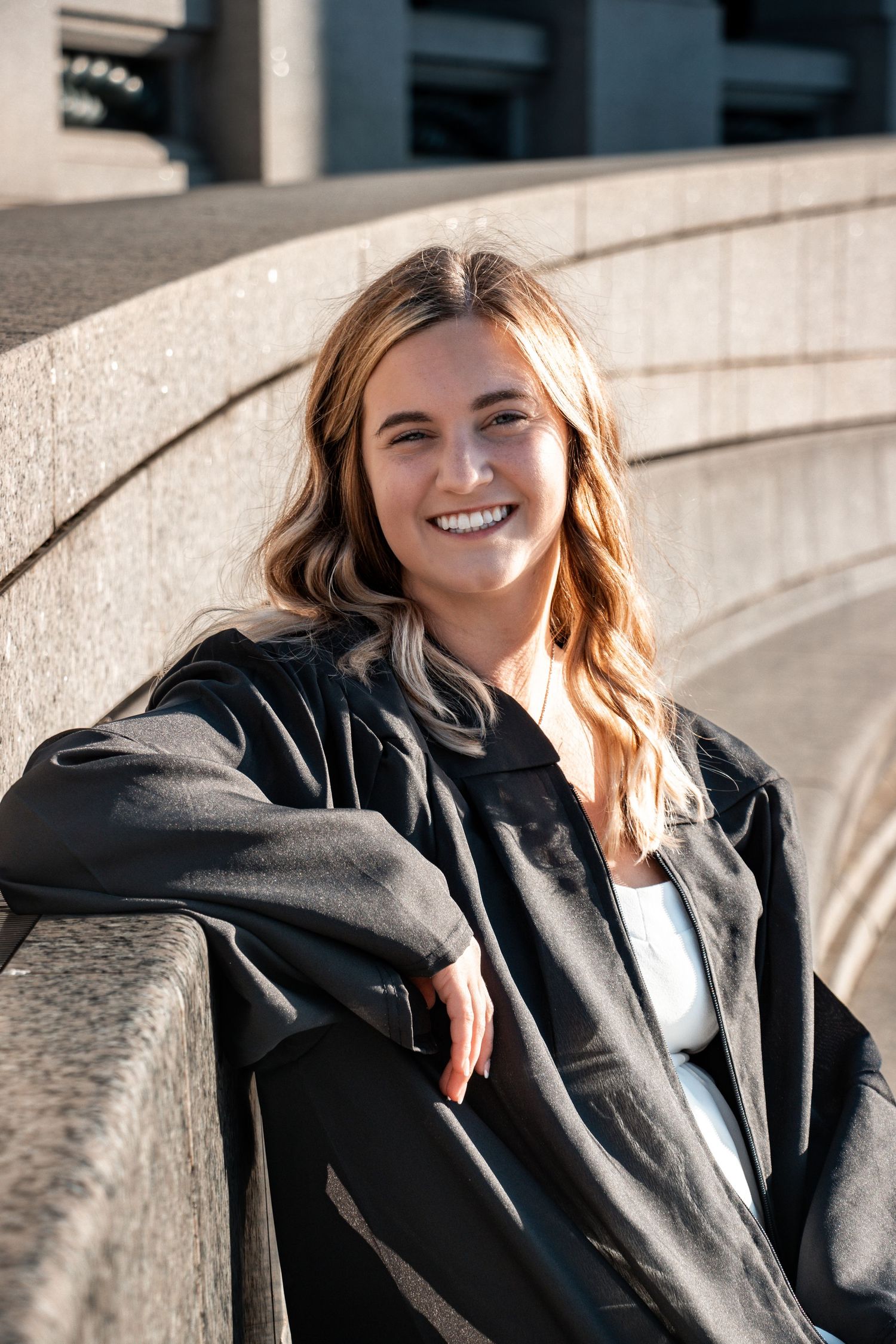 Graduate in black jacket sitting on concrete wall during sunny outdoor portrait session.