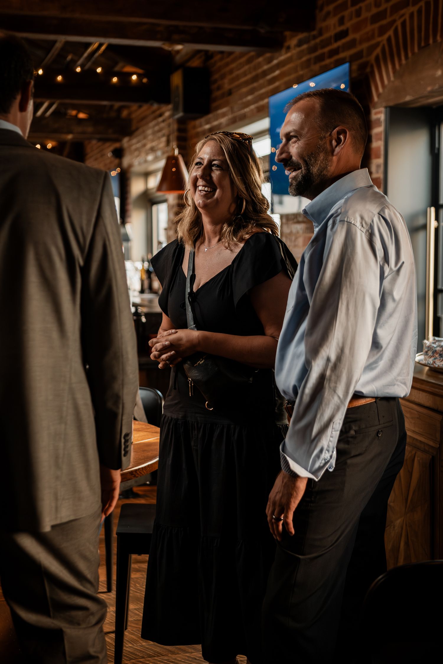 Event guests in formal attire socializing in a rustic venue with string lights overhead.